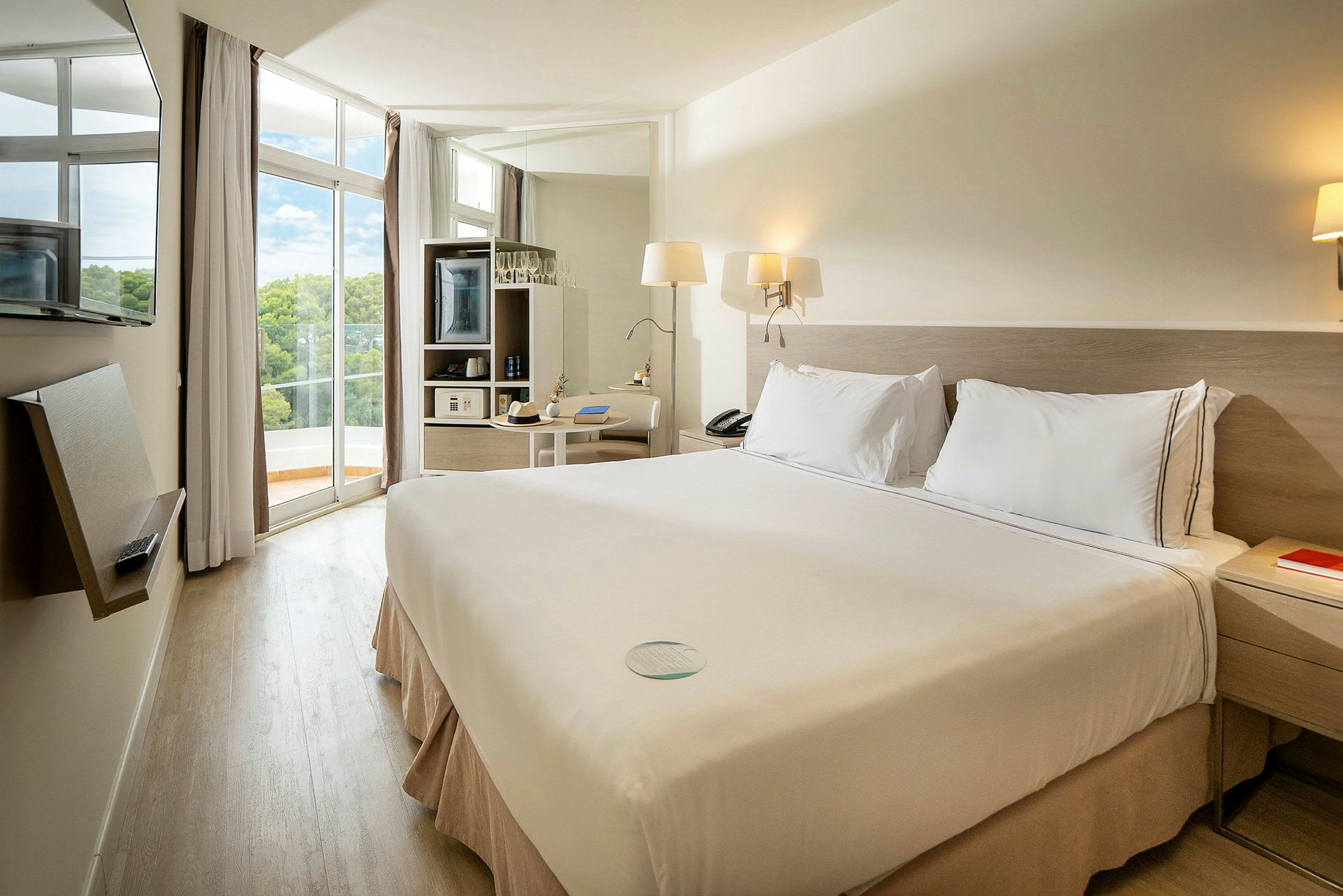 Hotellrom med dobbeltseng og lys innredning, store vinduer og balkong med utsikt. (AI-generert tekst)