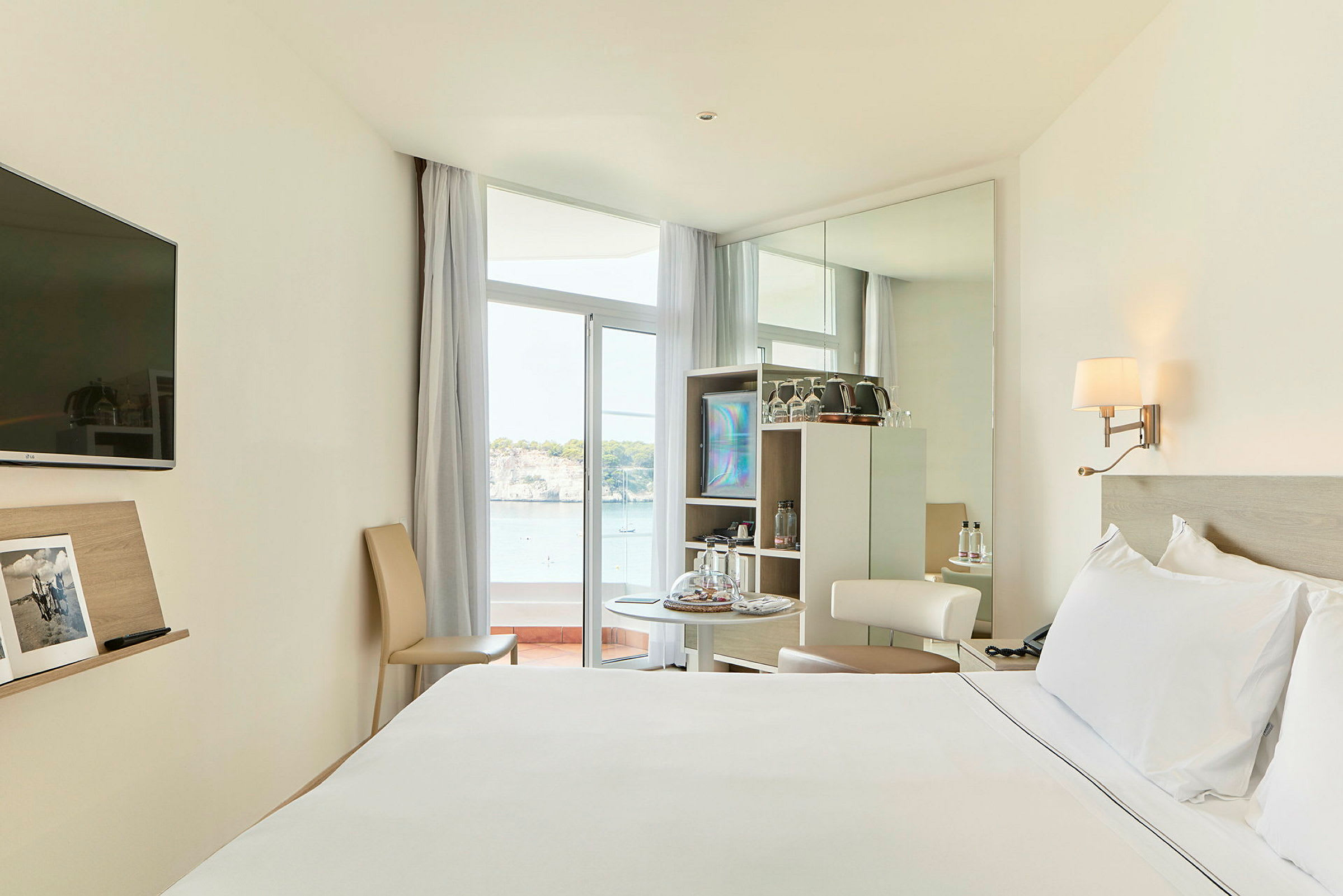 Et hotellrom med en stor seng, et lite bord med stoler, og utsikt mot havet gjennom en glassdør. (AI-generert tekst)