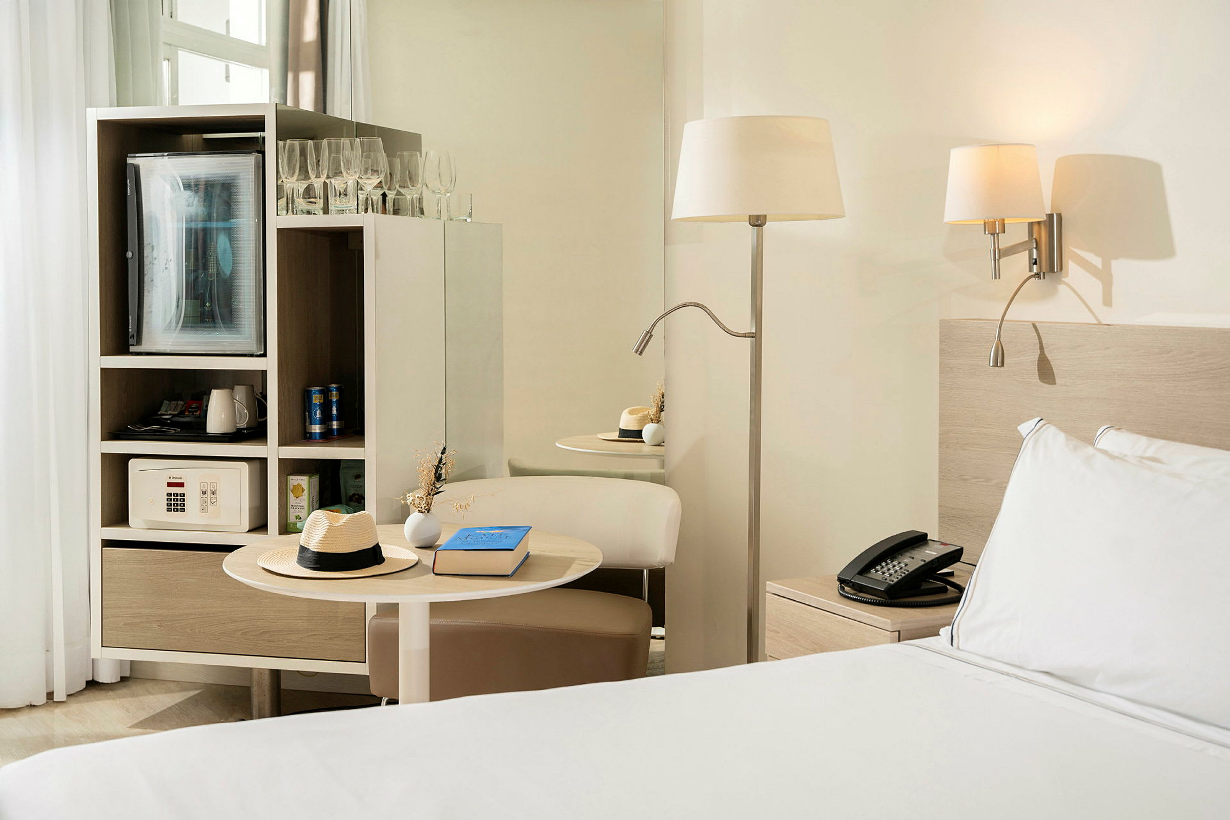 Et hotellrom med en seng ved siden av et nattbord, en bordslampe og en integrert møbel som inneholder et kjøleskap og en safe. (AI-generert tekst)