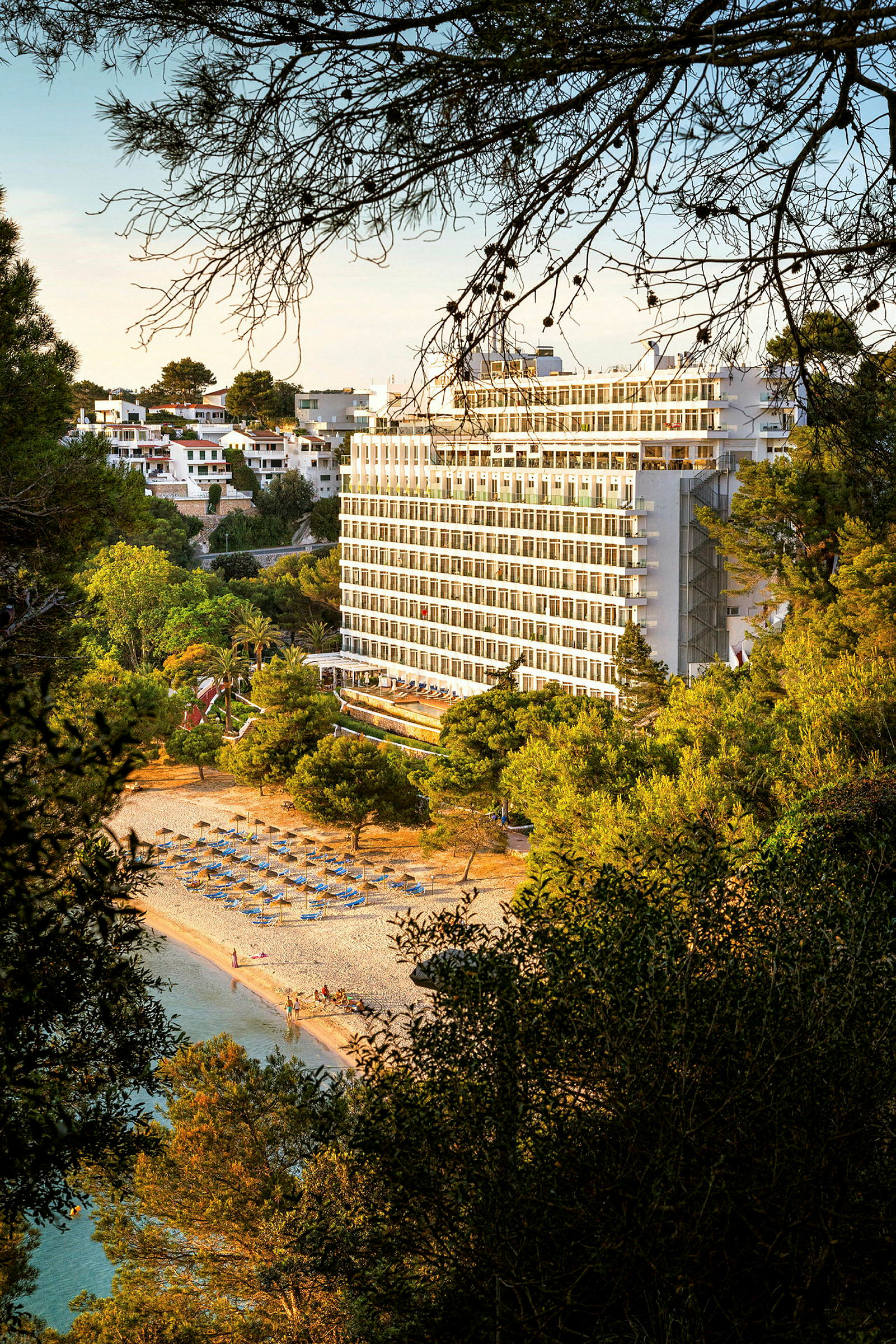Bilde av et hotell bygget nær en strand med solsenger og parasoller, omgitt av frodige trær. (AI-generert tekst)