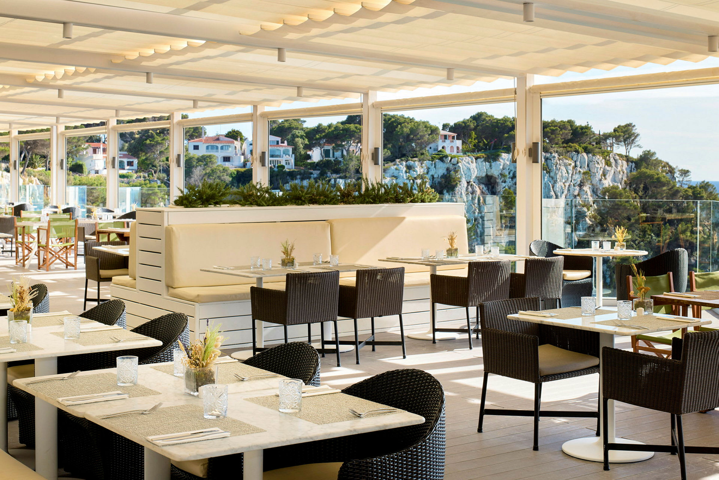 En solrik restaurantterrasse med bord og stoler samt utsikt over et frodig område med hus på en forhøyning. (AI-generert tekst)