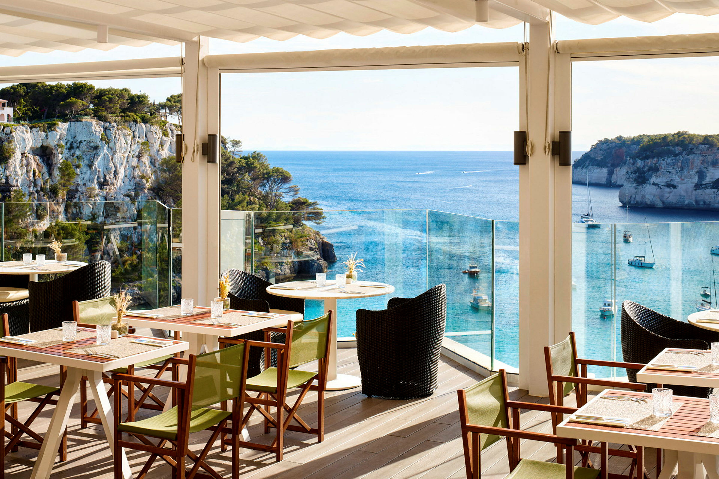 En restaurantterrasse med bord og stoler, med utsikt over havet og klipper. (AI-generert tekst)
