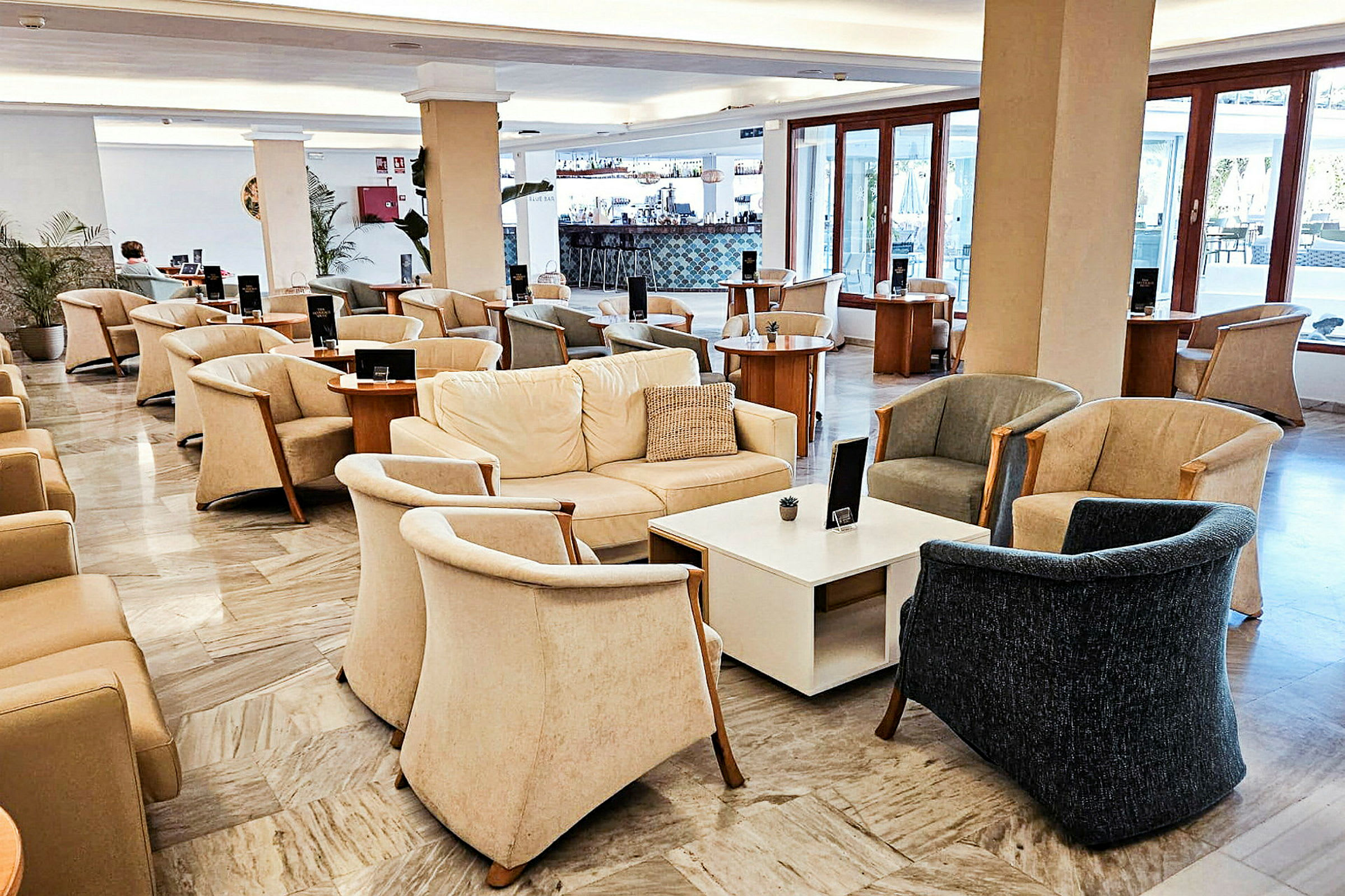 En lounge med komfortable lenestoler, sofaer og bord, med store vinduer i bakgrunnen. (AI-generert tekst)