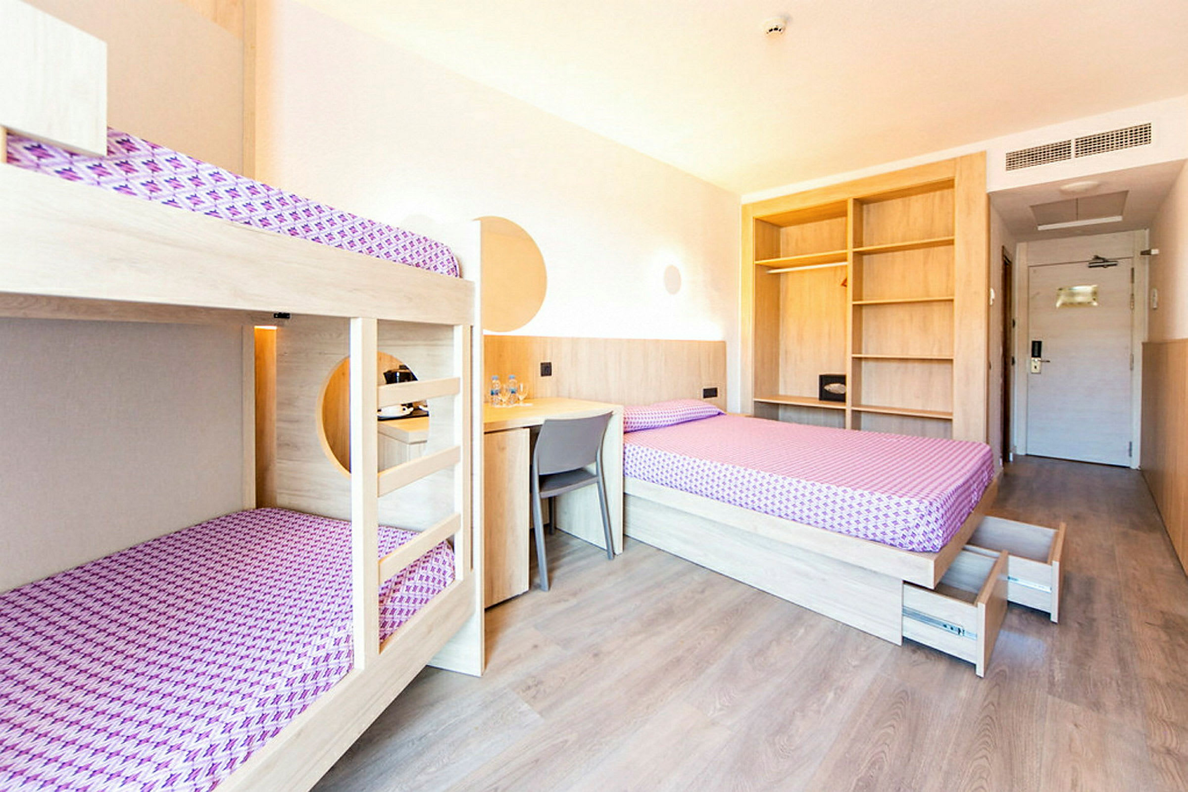 Et hotellrom med køyesenger, en enkeltseng, skrivebord og åpne hyller. Et dørområde kan ses i bakgrunnen. (AI-generert tekst)
