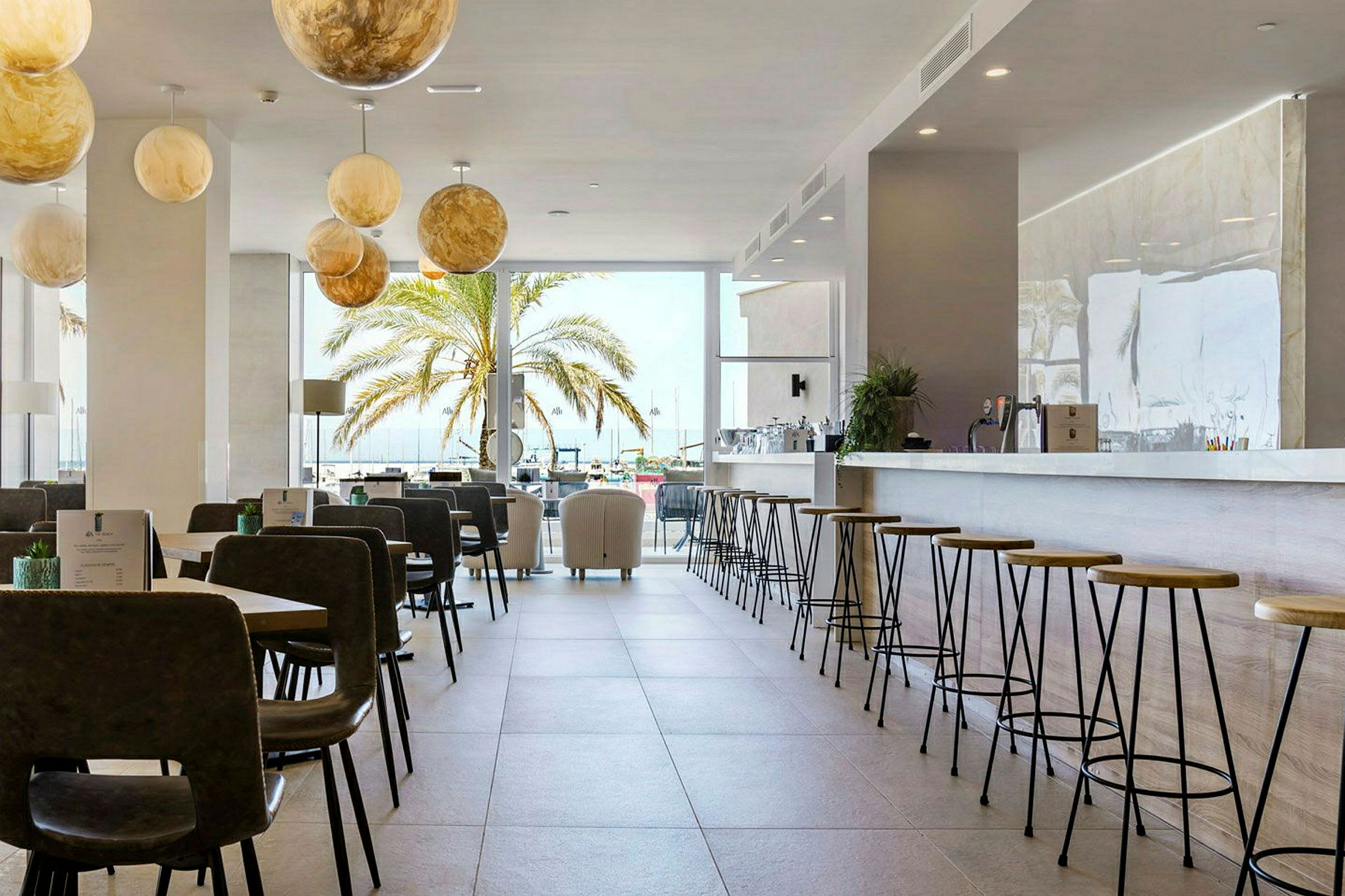 En lys bar og restaurant med sitteplasser, samt en palme utenfor et stort vindu. (AI-generert tekst)