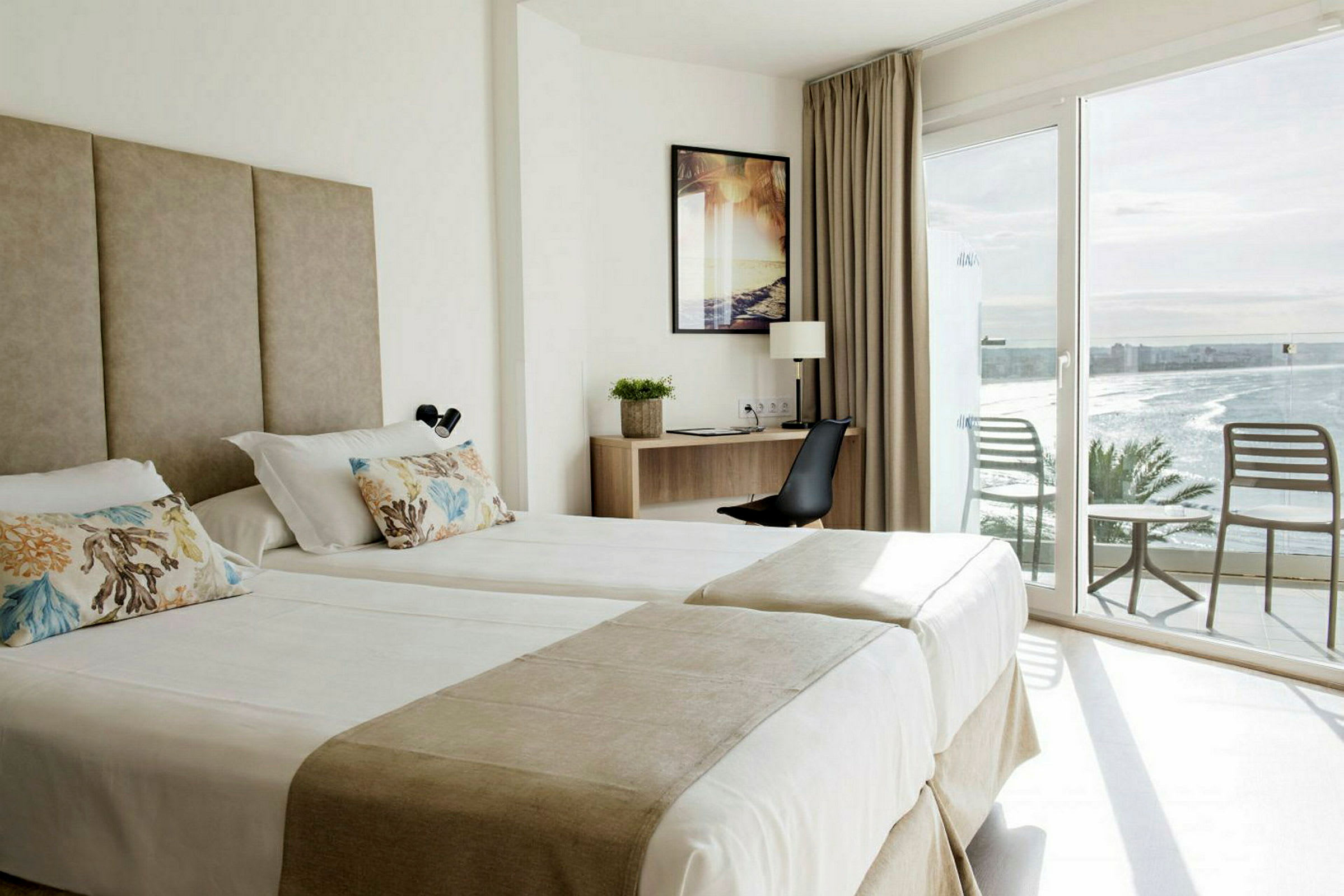 Hotellrom med to enkeltsenger, skrivebord og to stoler på balkongen med havutsikt. (AI-generert tekst)