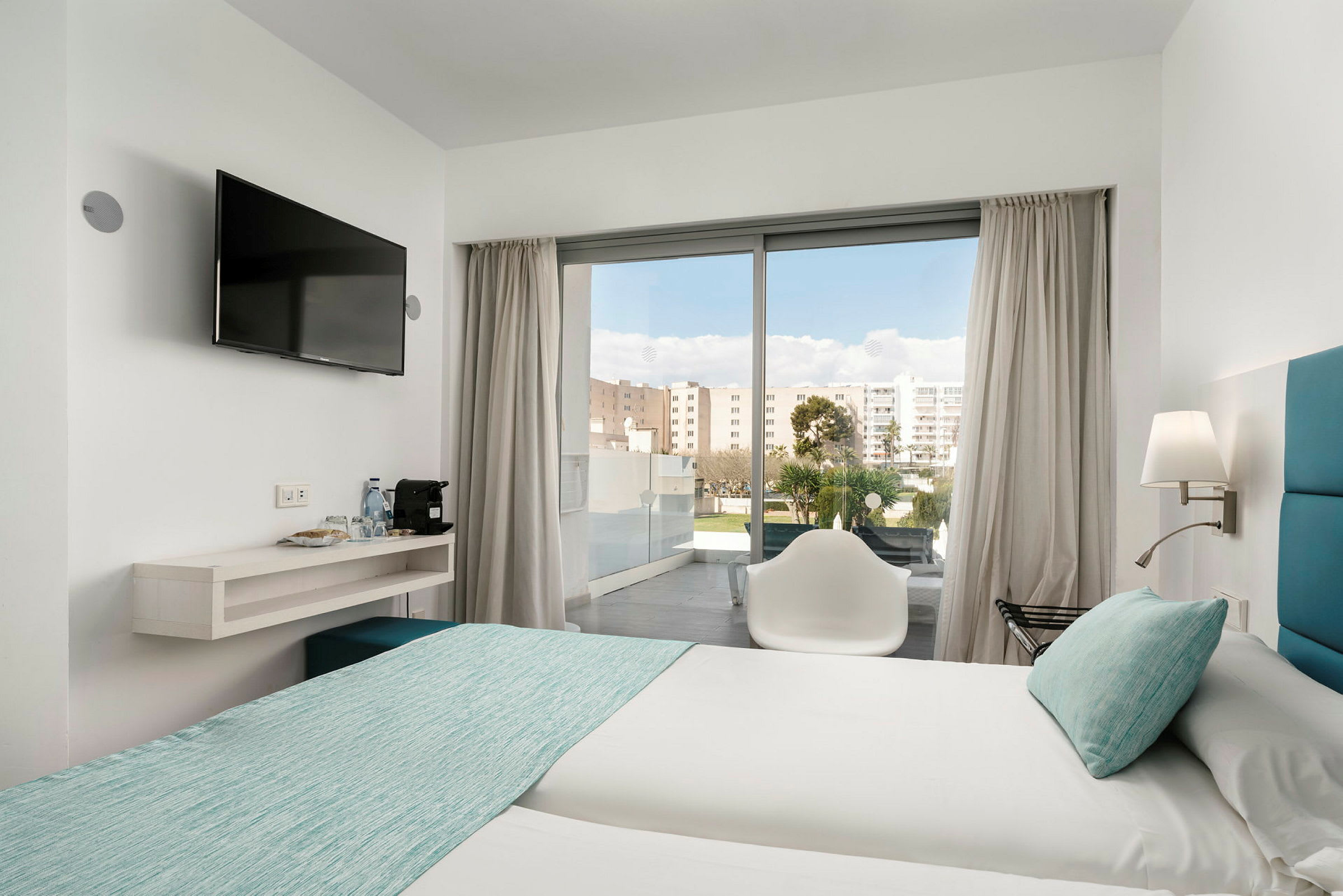 Et hotellrom med to senger oppredd med lyse laken og turkise sengetepp. En stol står foran en glassdør som leder ut til en balkong. En veggmontert TV henger ved siden av en hylle med vannflasker og kaffemaskin. (AI-generert tekst)