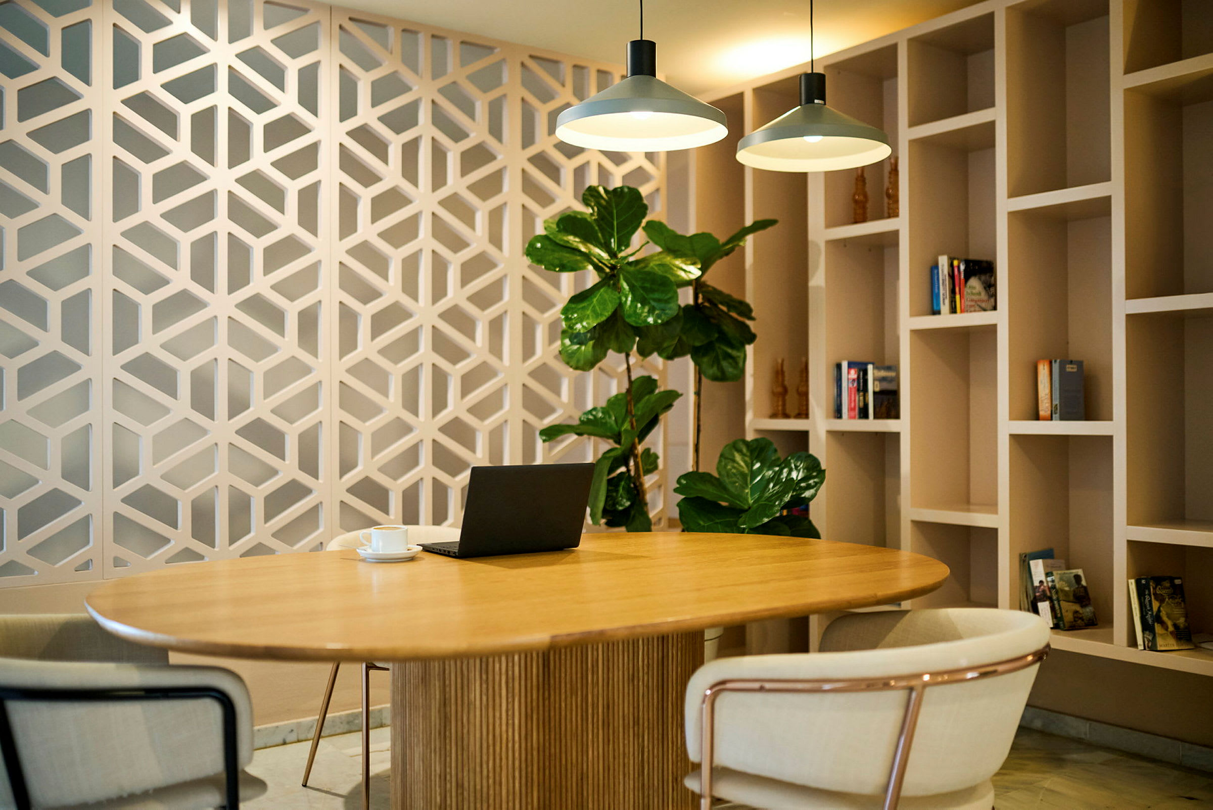 Et moderne og stilrent arbeidsområde med et rundt trebord, omkringliggende stoler, plantepynt og en vegg med hyller og geometriske detaljer. (AI-generert tekst)