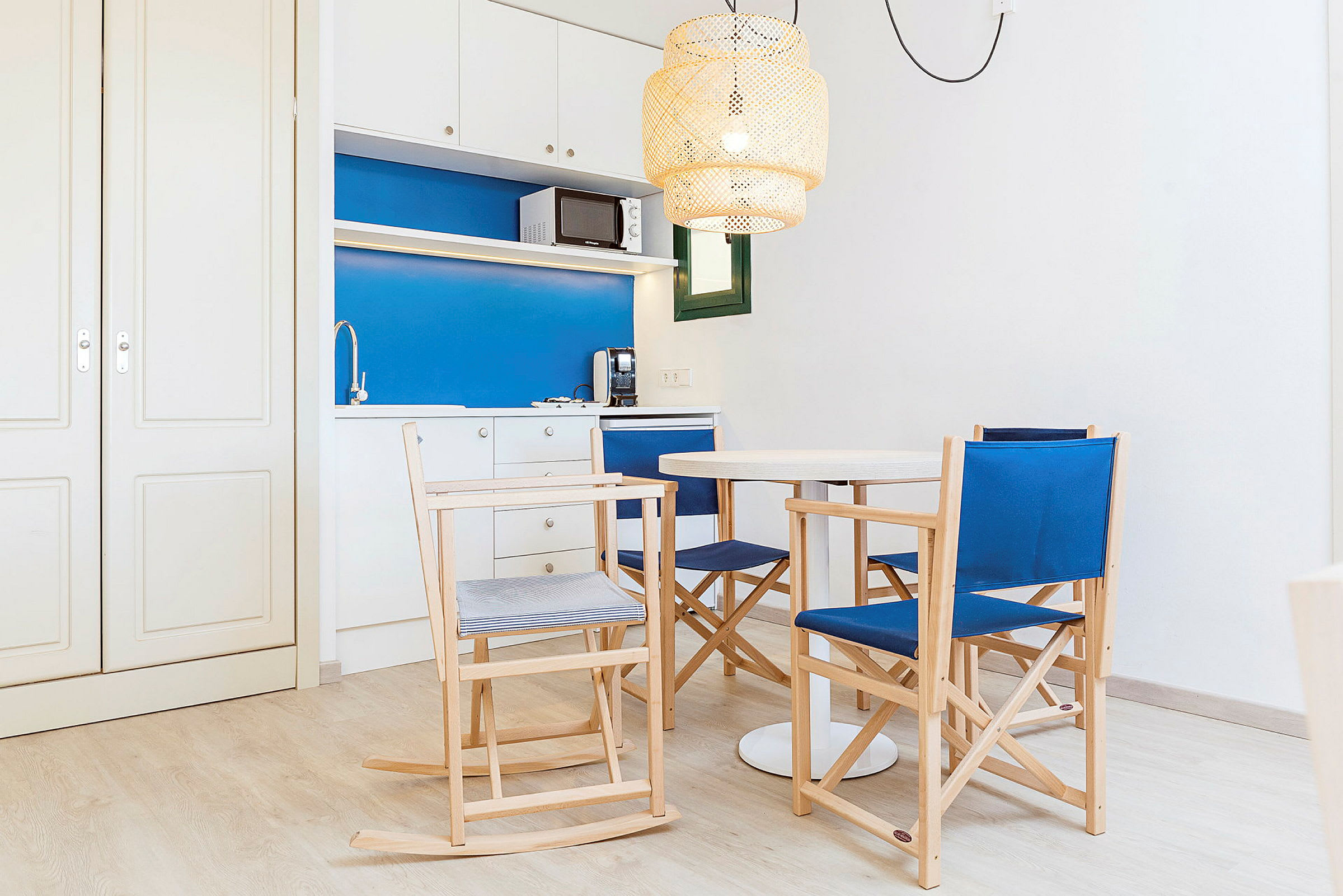 Et lite kjøkken med blå og hvite flater, en spisegruppe med trestoler og et rundt hvitt bord. En taklampe med flettet design henger over bordet. (AI-generert tekst)