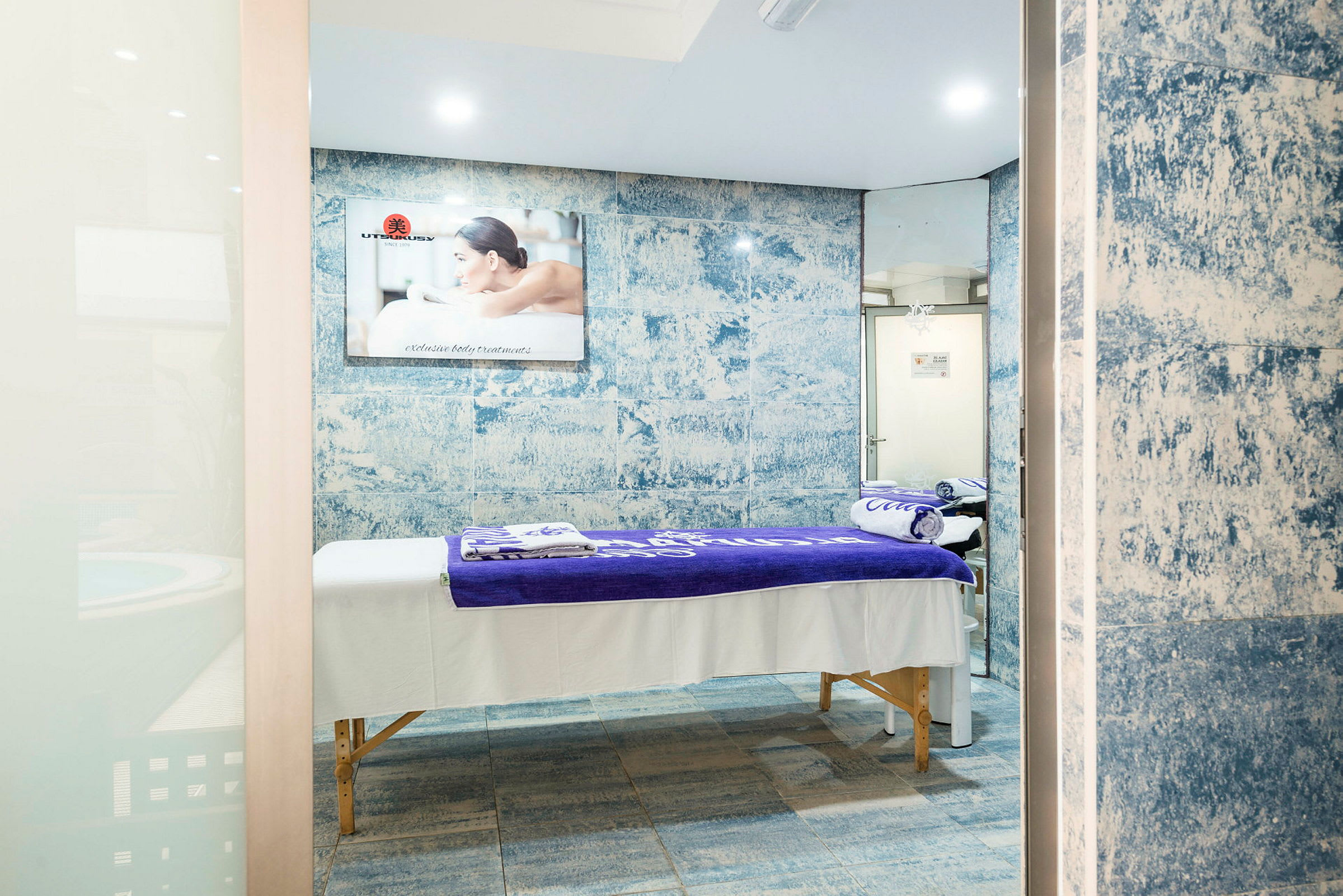Et spa-rom med massasjebenk dekket av hvite og lilla håndklær, blå mosaikk veggdesign og speil. (AI-generert tekst)