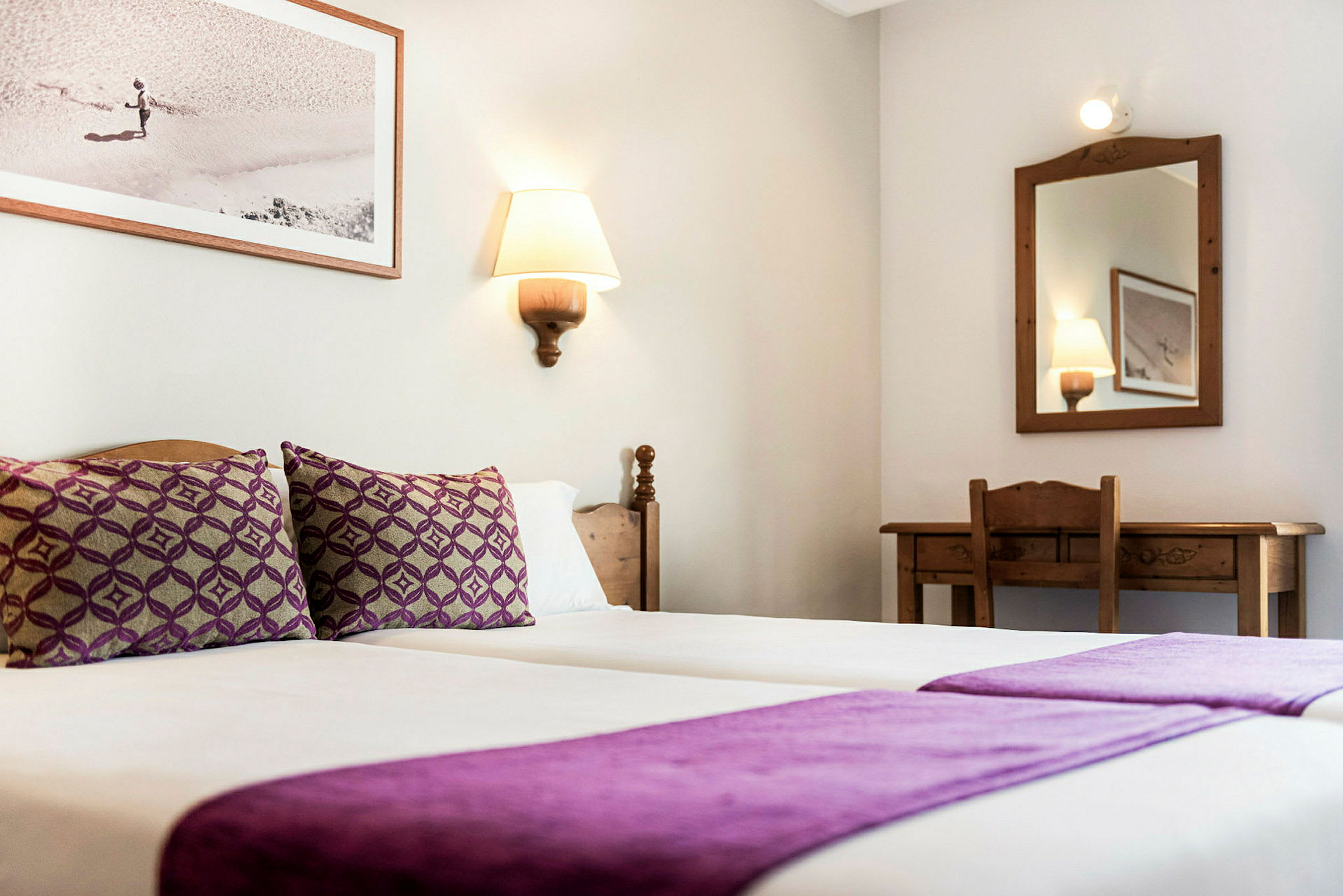 Et hotellrom med to enkeltsenger med lilla sengetøy, et speil og et skrivebord. (AI-generert tekst)