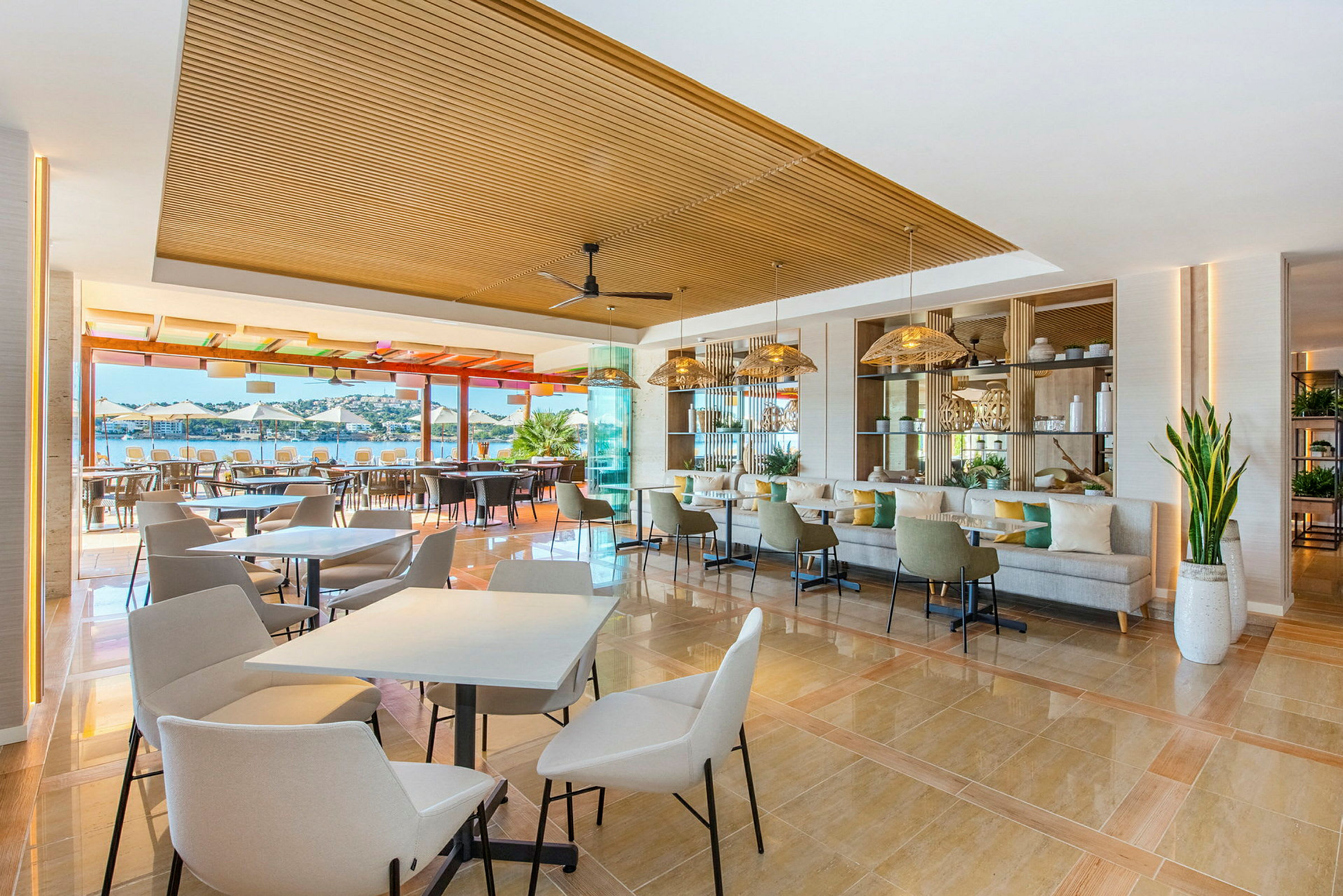 Et moderne og åpent restaurantområde med bord og stoler, innredet i lyse farger og store vinduer som vender ut mot en utendørsterrasse og havutsikt. (AI-generert tekst)