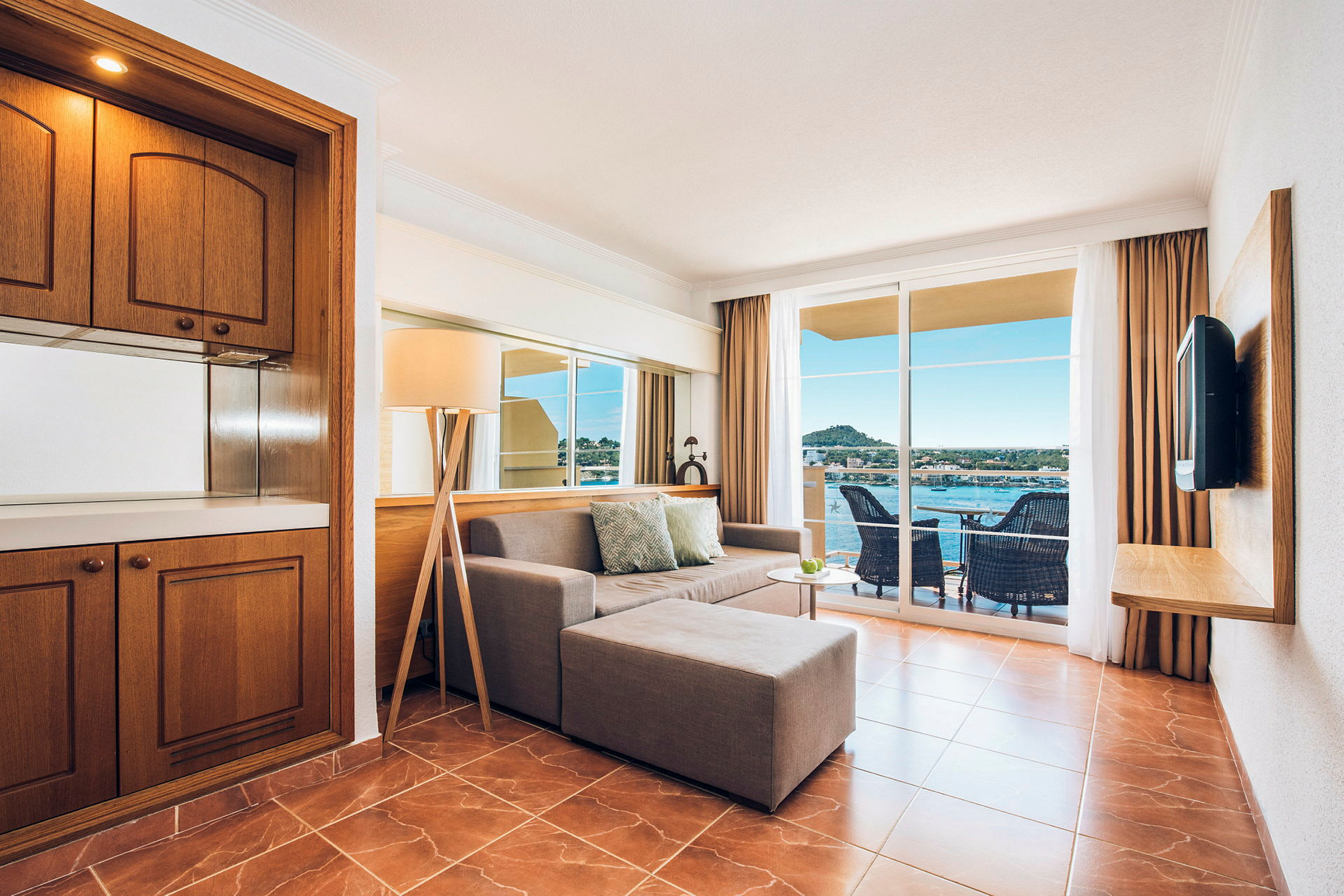 Et hotellrom med sofa, sofabord og brunt gulv. Store vinduer med utsikt over balkongen og havet. (AI-generert tekst)