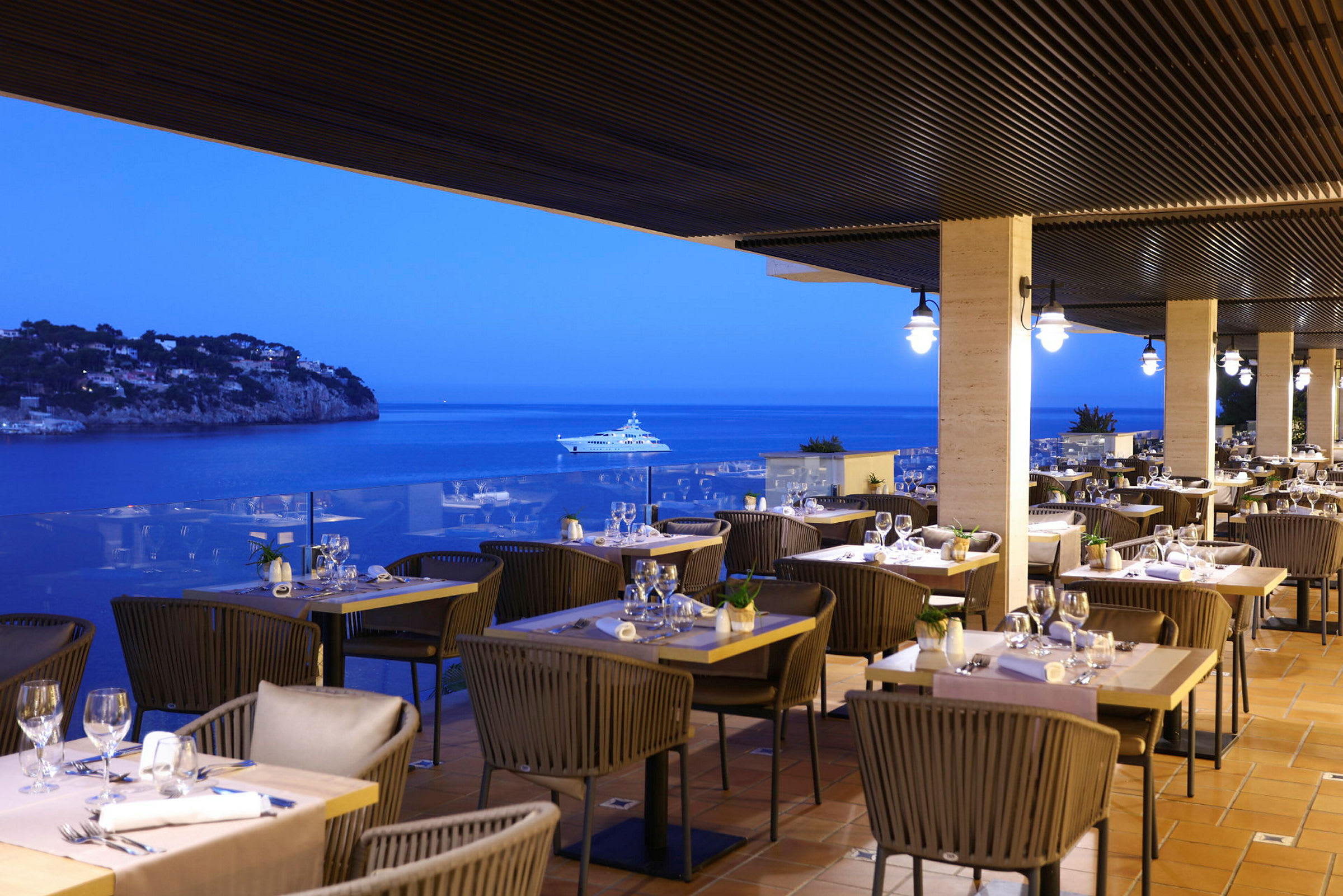 En restaurant med uteservering på en terrasse med utsikt over havet om kvelden. Flere bord er dekket med stoler og glass. (AI-generert tekst)