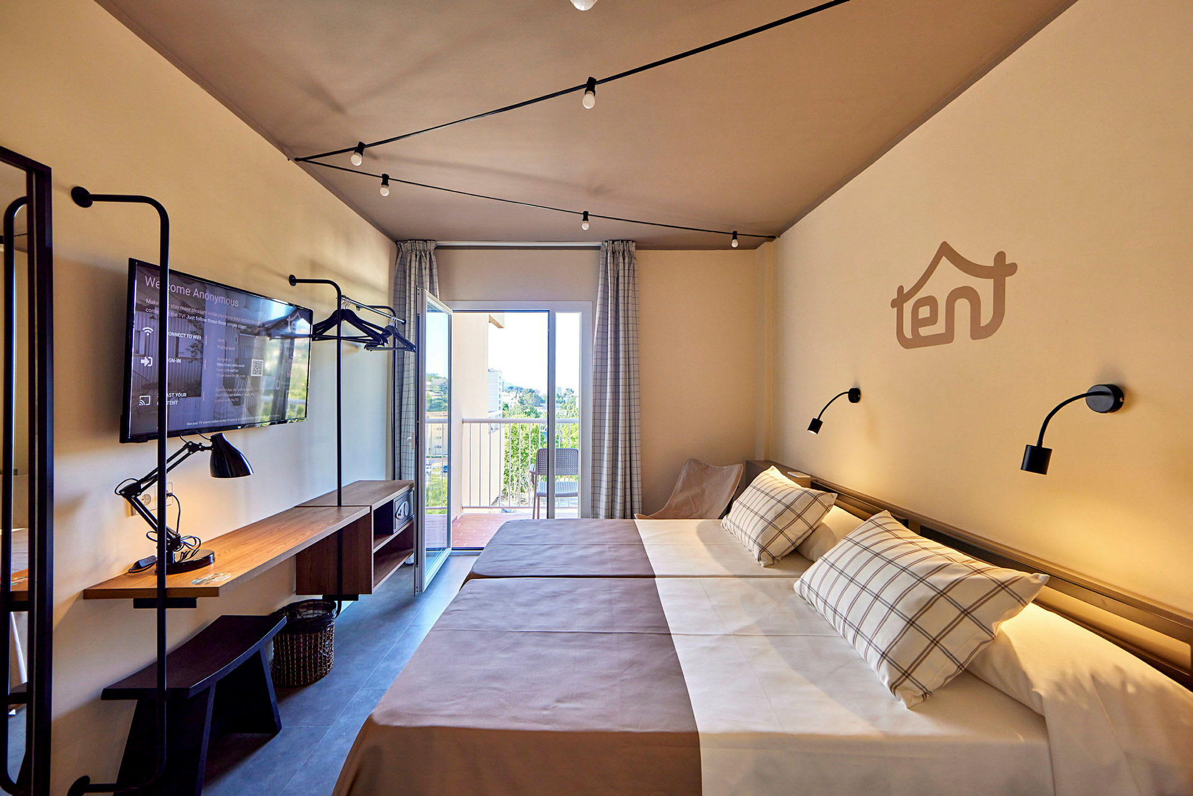 Hotellrom med to enkeltsenger, arbeidsbord med lampe og TV, samt balkong med utsikt. (AI-generert tekst)