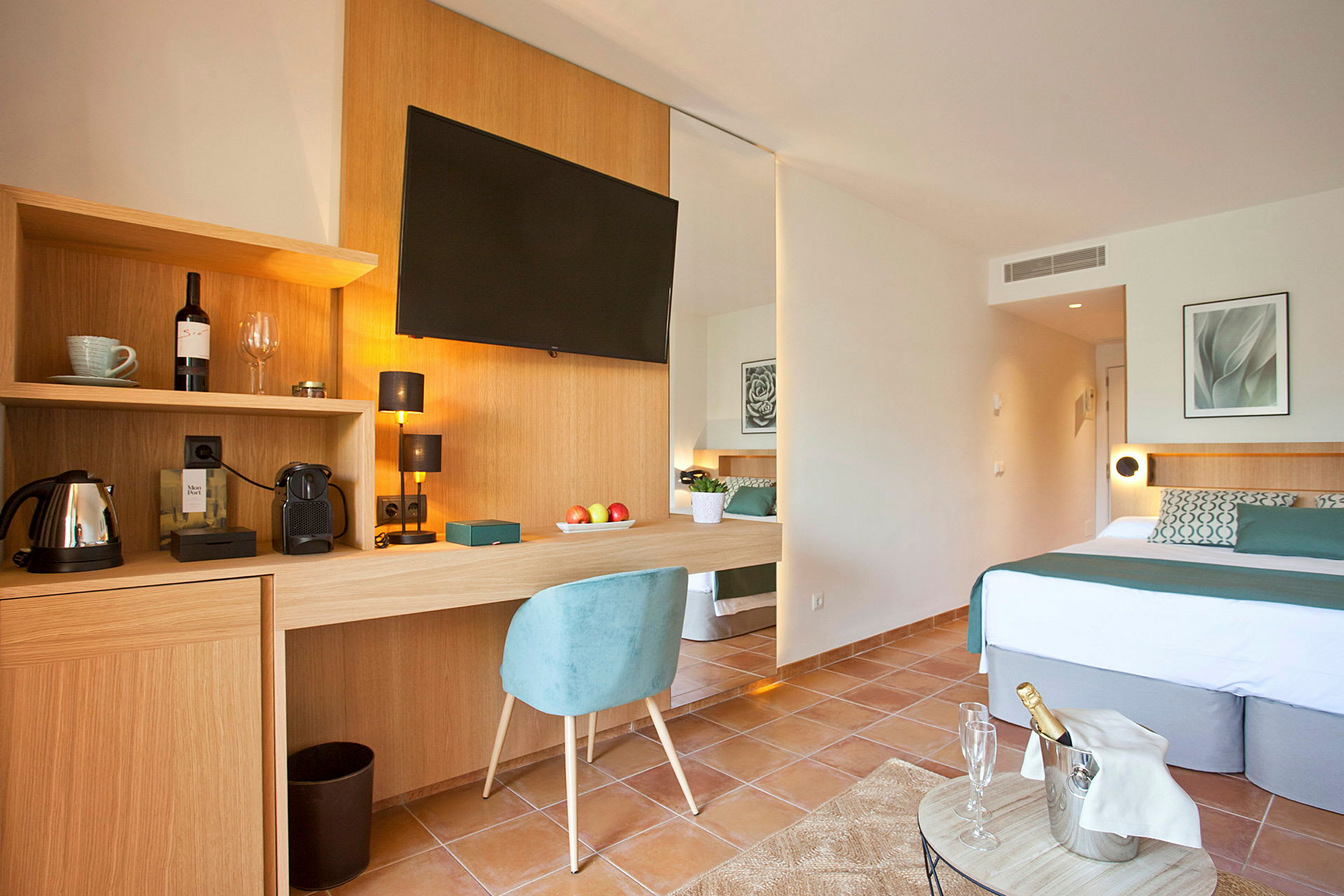 Et moderne hotellrom med en dobbeltseng, en skrivebordsflate med stol, en veggmontert TV og inkludert kaffemaskin. Dekorasjonen inkluderer et fotografi og en vinflaske, samt en fylt champagnekjøler på et lite bord. (AI-generert tekst)