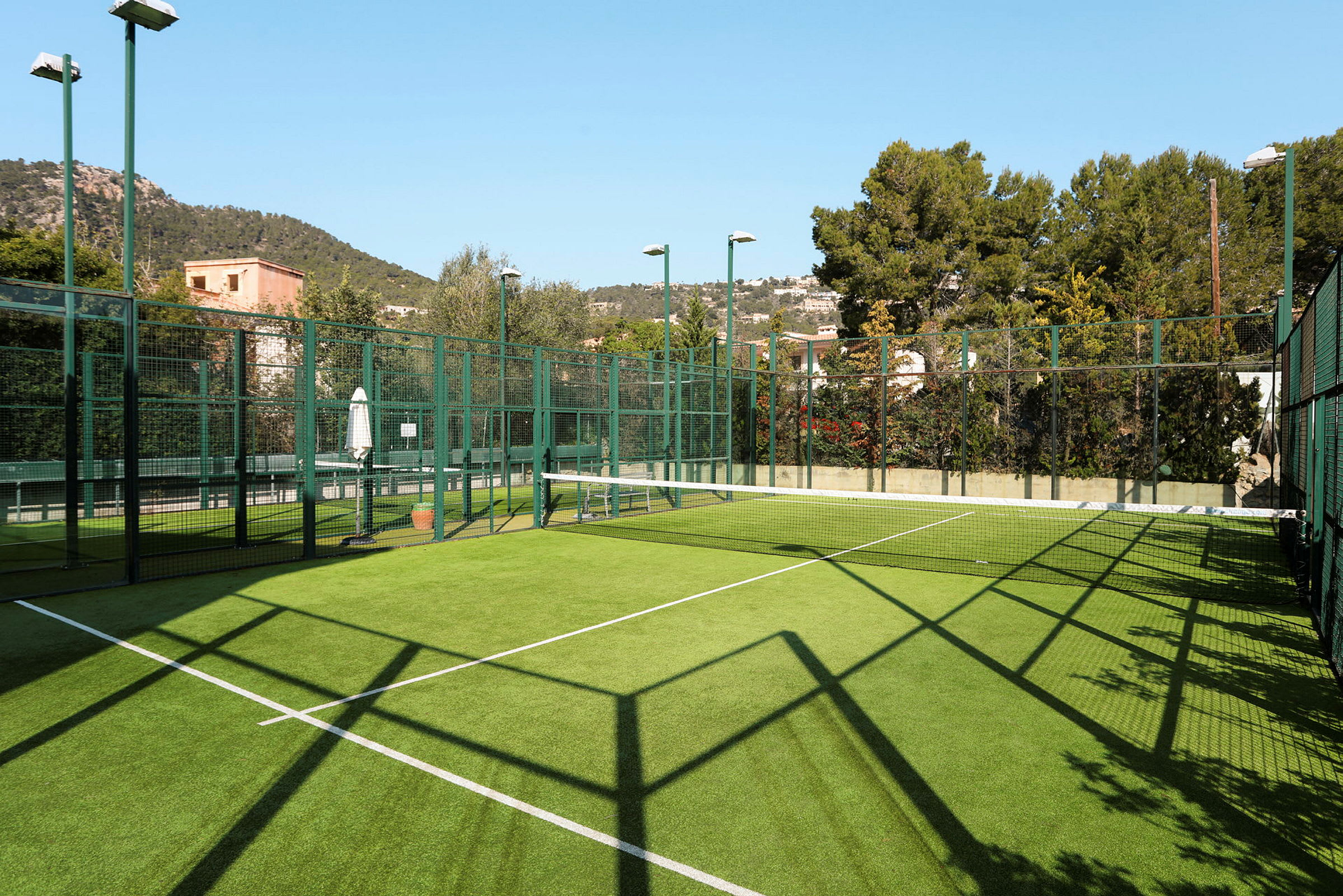 En padelbane utendørs med grønn kunstgressoverflate, omgitt av beskyttelsesnett og trær og bygninger. (AI-generert tekst)