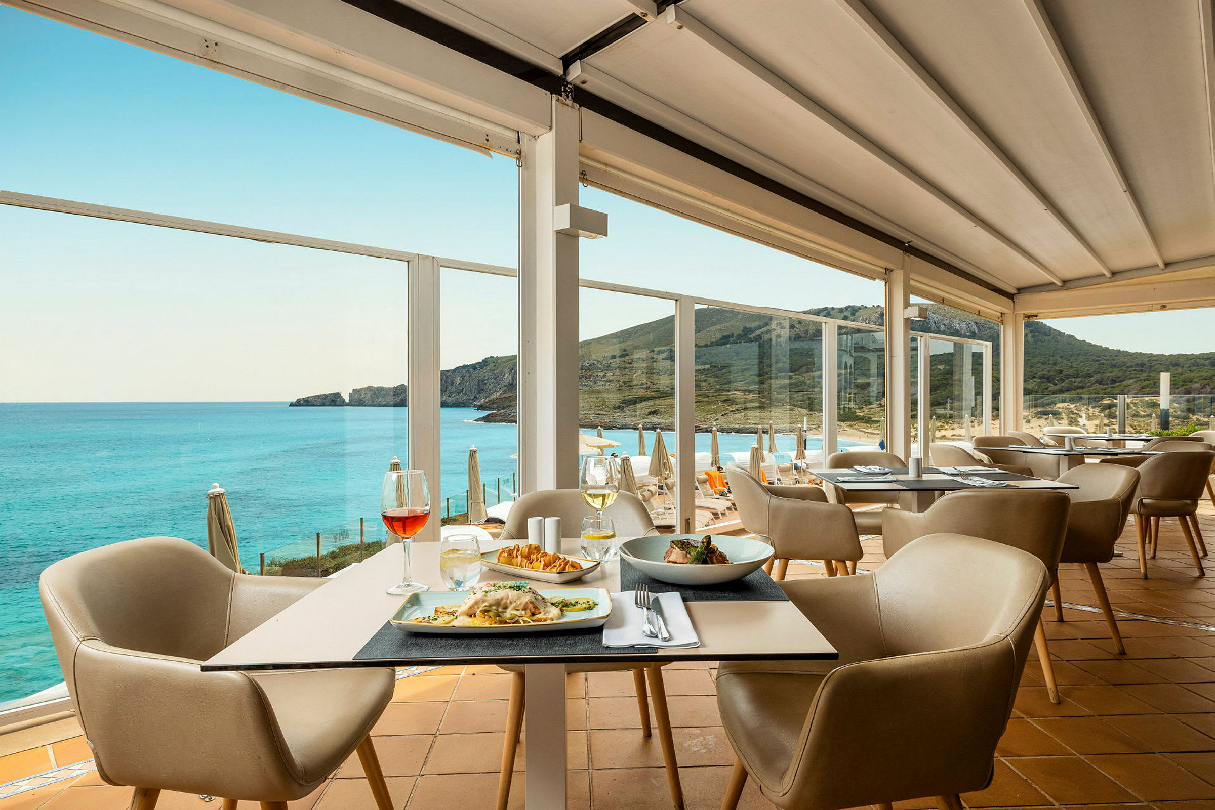 En restaurant med bord med mat og stoler, samt en panoramautsikt over havet og en fjellkyst. (AI-generert tekst)
