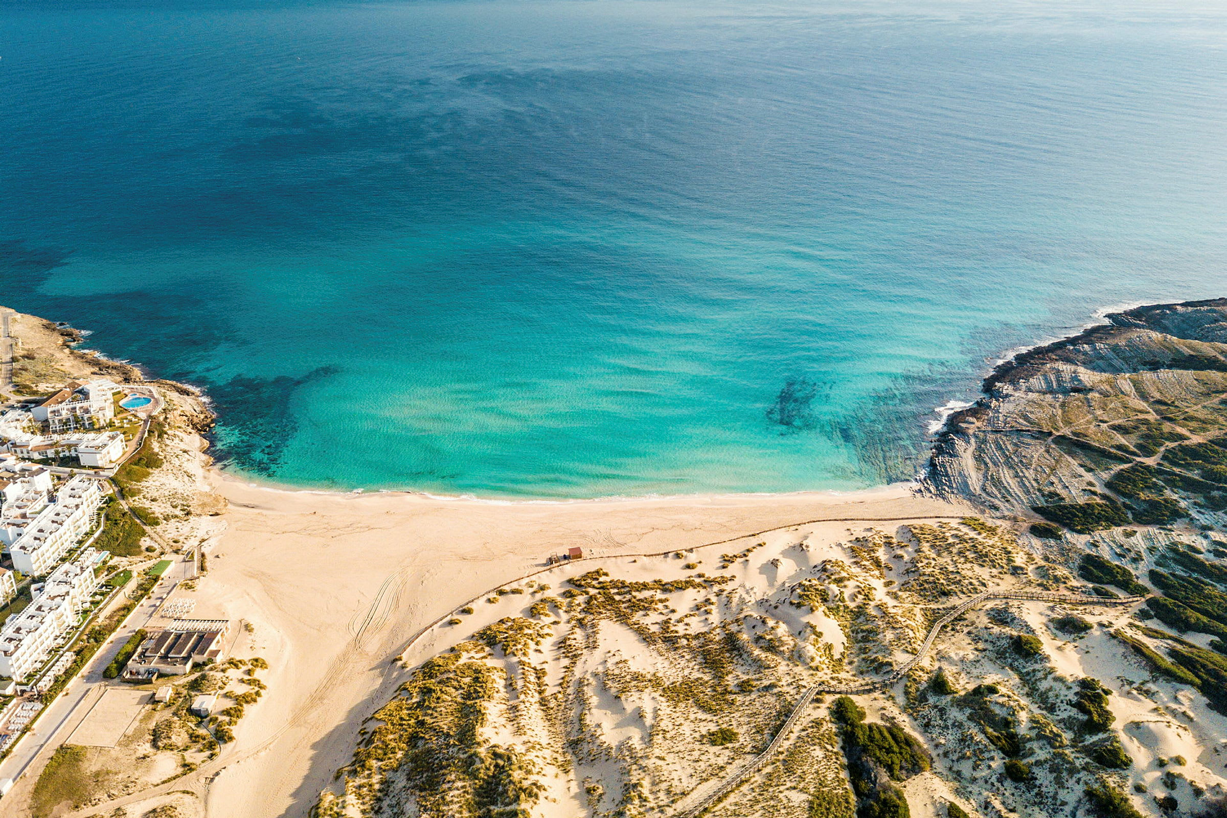 Flyfoto av en strand med turkist vann og sanddyner, omgitt av kystgrønne områder. (AI-generert tekst)