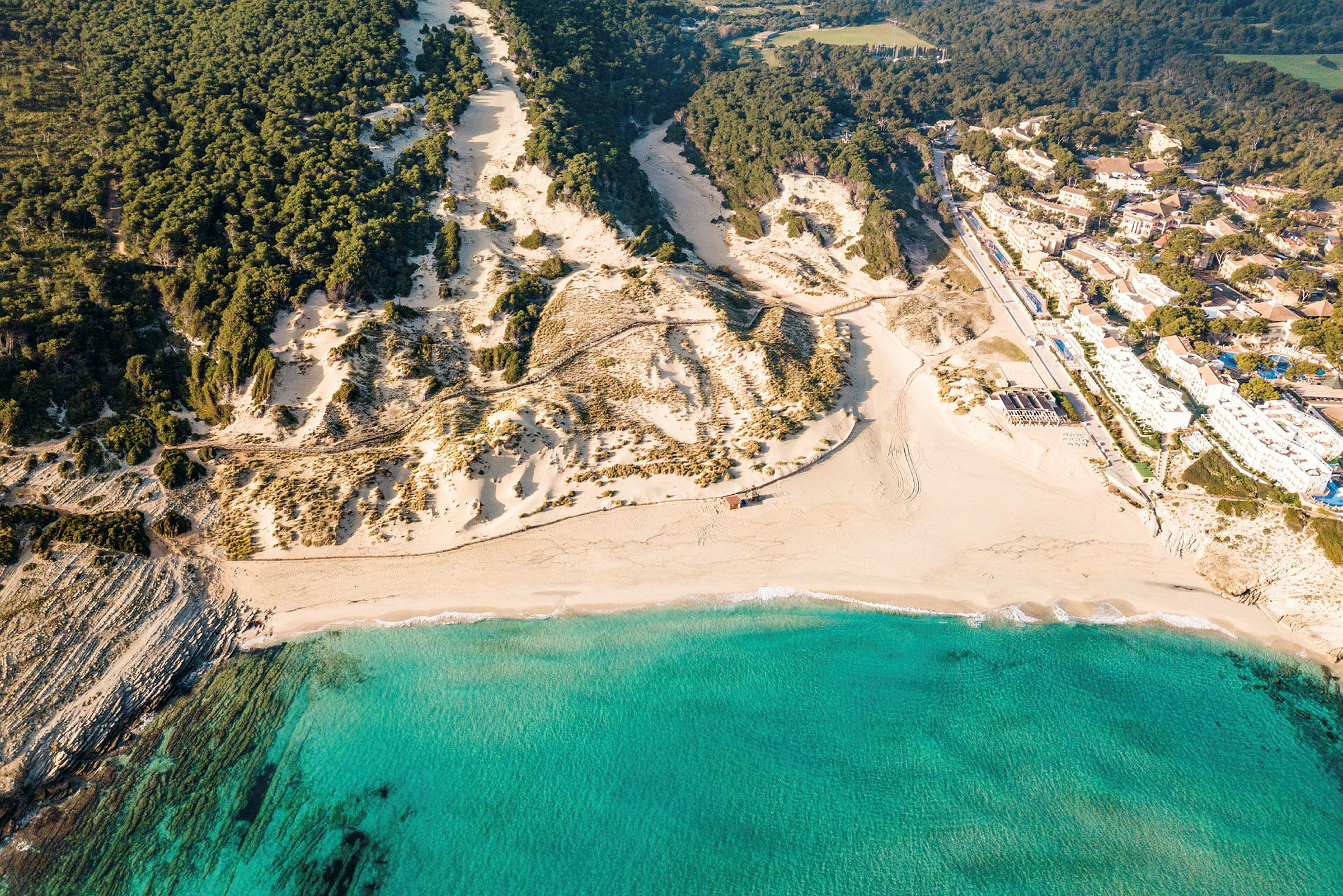 Flyfoto av en sandstrand med turkis vann, omgitt av klipper og furuskog. (AI-generert tekst)