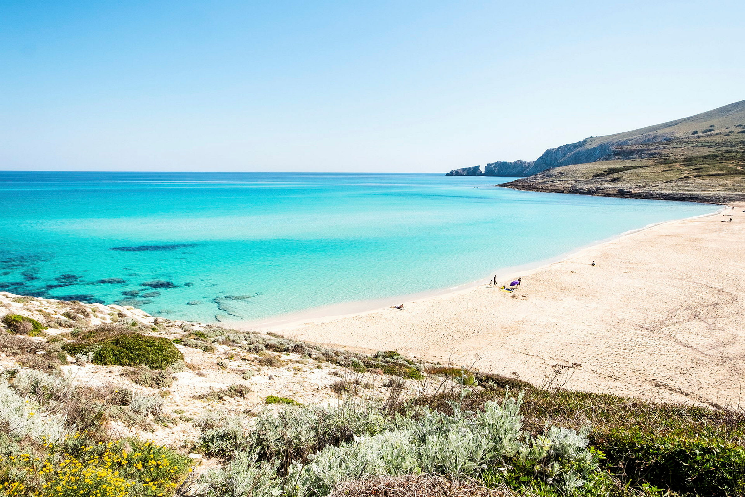 En strand med lys sand og klart blått vann, omgitt av grøntområder. (AI-generert tekst)