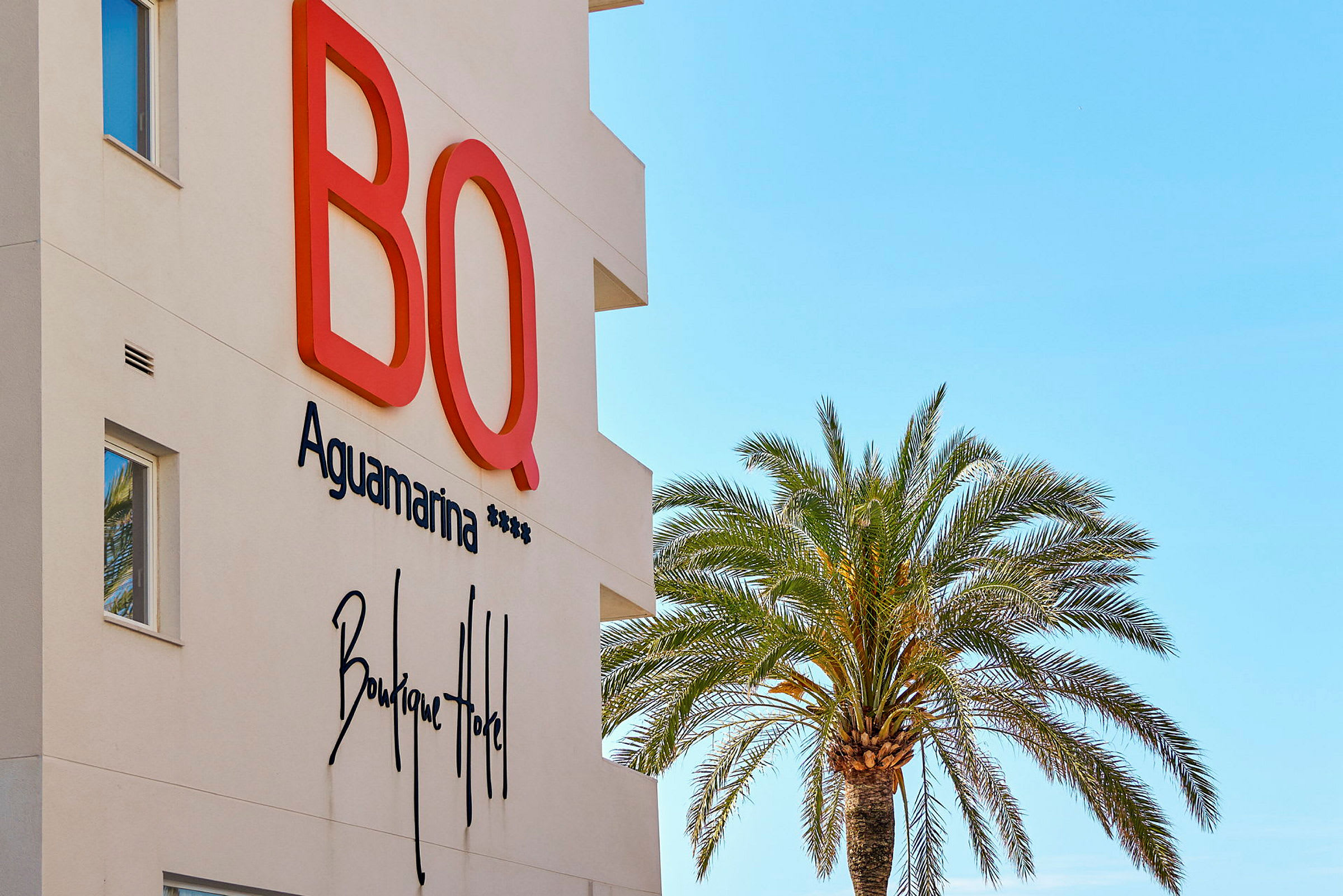 Fasaden til BQ Aguamarina Boutique Hotel med en palme mot klar blå himmel. (AI-generert tekst)