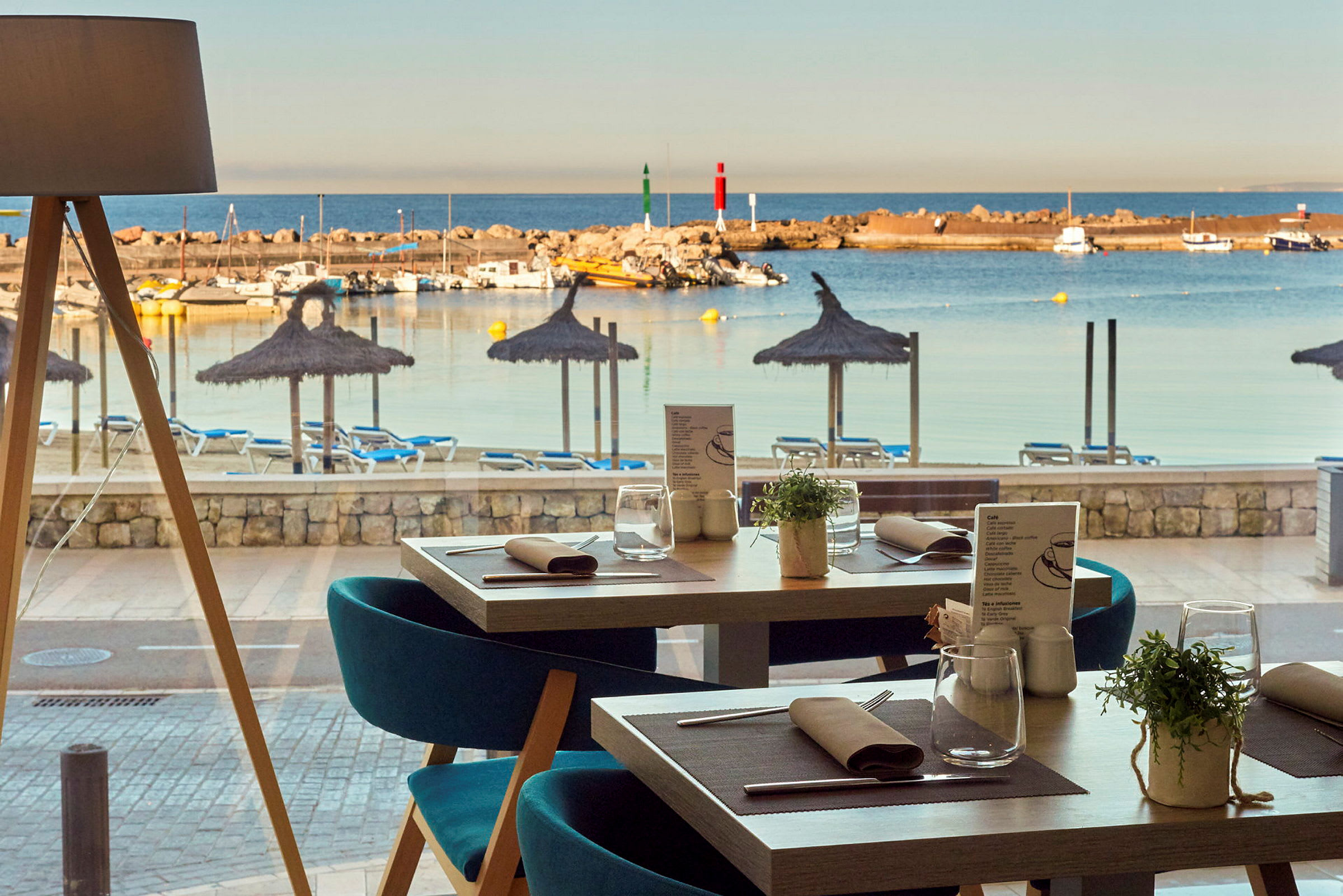 Utsikt fra en restaurant mot en strand med parasoller og solsenger, samt en småbåthavn med båter. (AI-generert tekst)