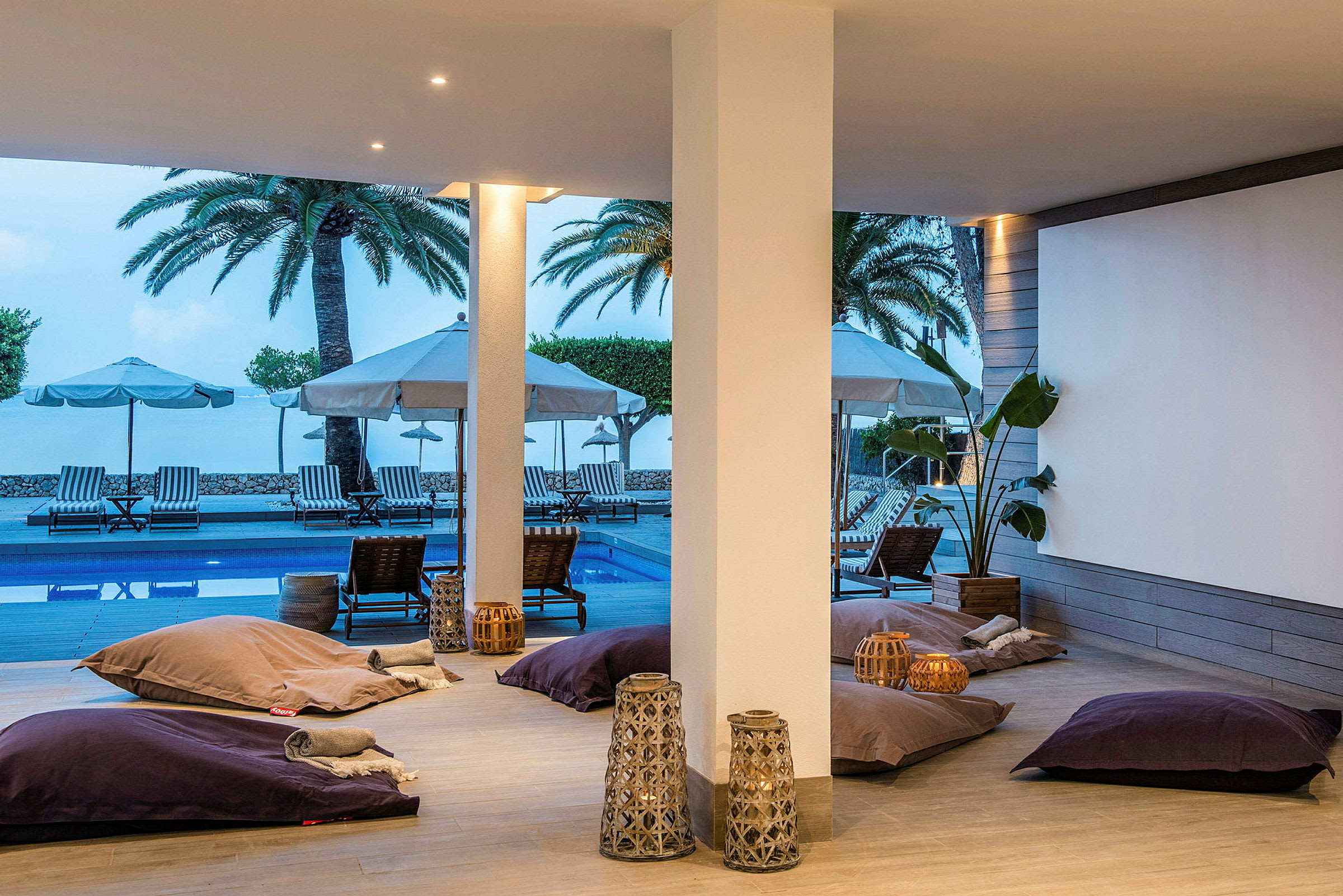 En åpen lounge med store puter og dekorative lyslykter. I bakgrunnen sees et bassengområde med solsenger og parasoller, samt palmer. (AI-generert tekst)