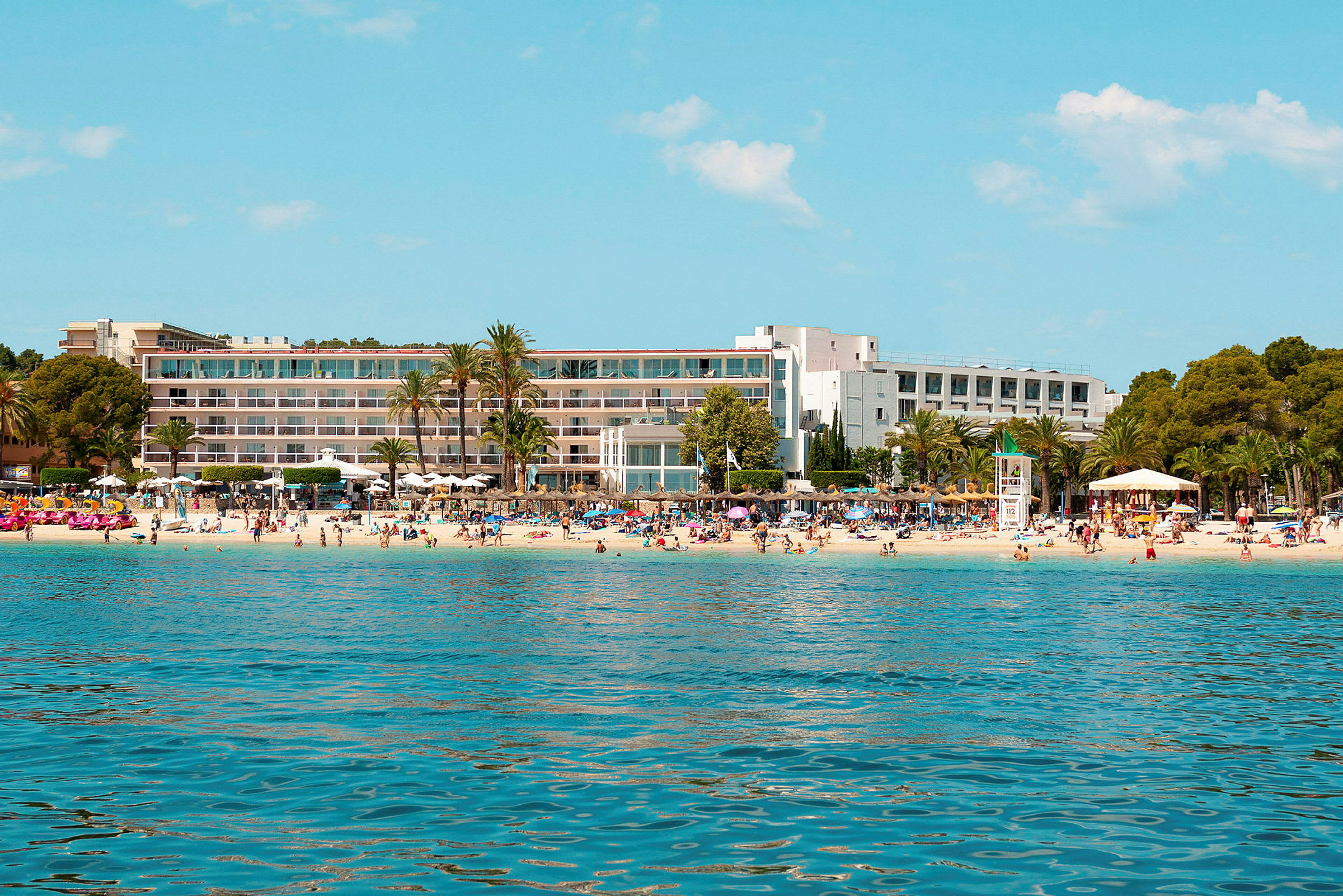 Et hotell ved en strand med palmer og badende personer. (AI-generert tekst)
