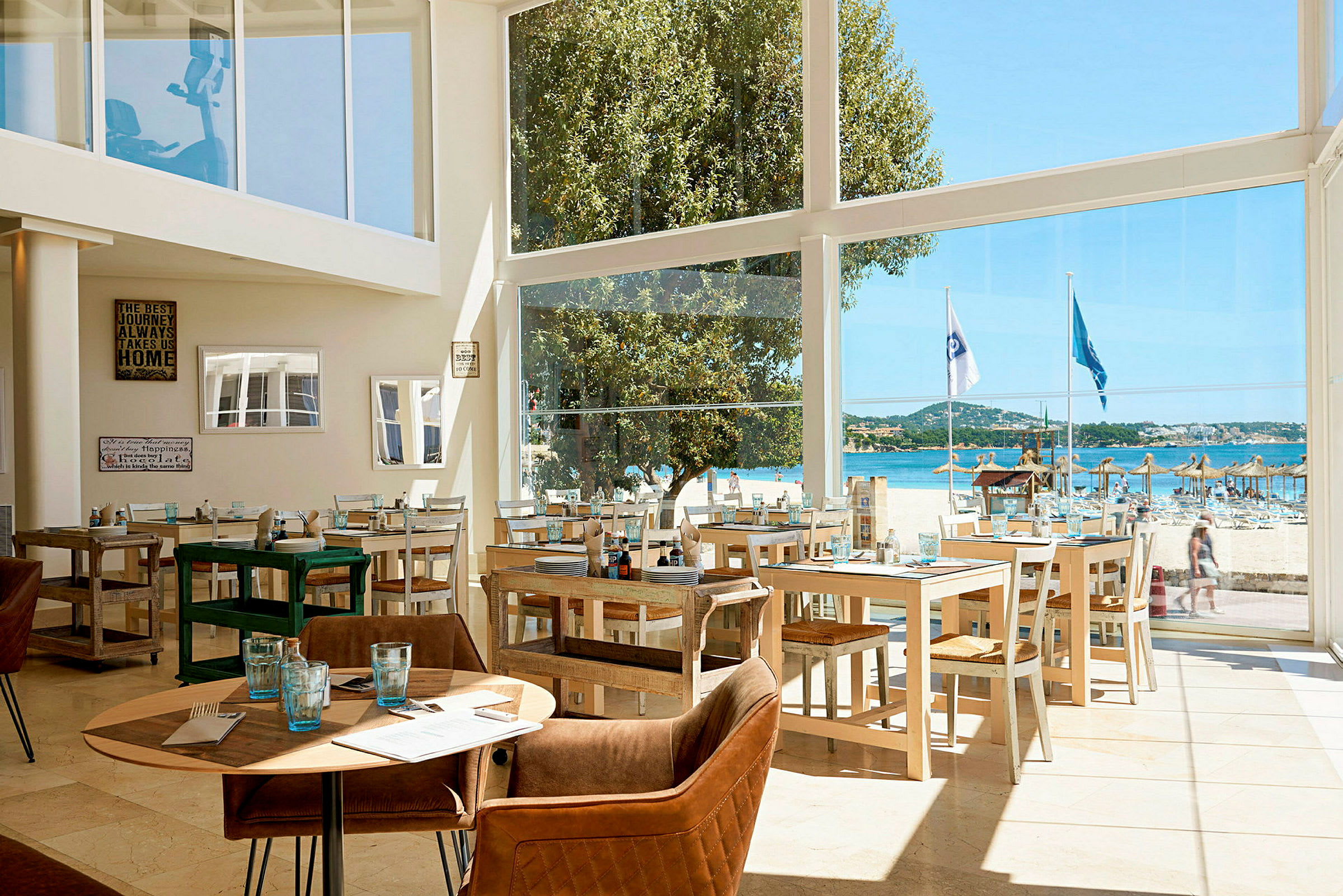 En restaurant med glassvegger som gir utsikt mot en strand med parasoller og blått hav. (AI-generert tekst)
