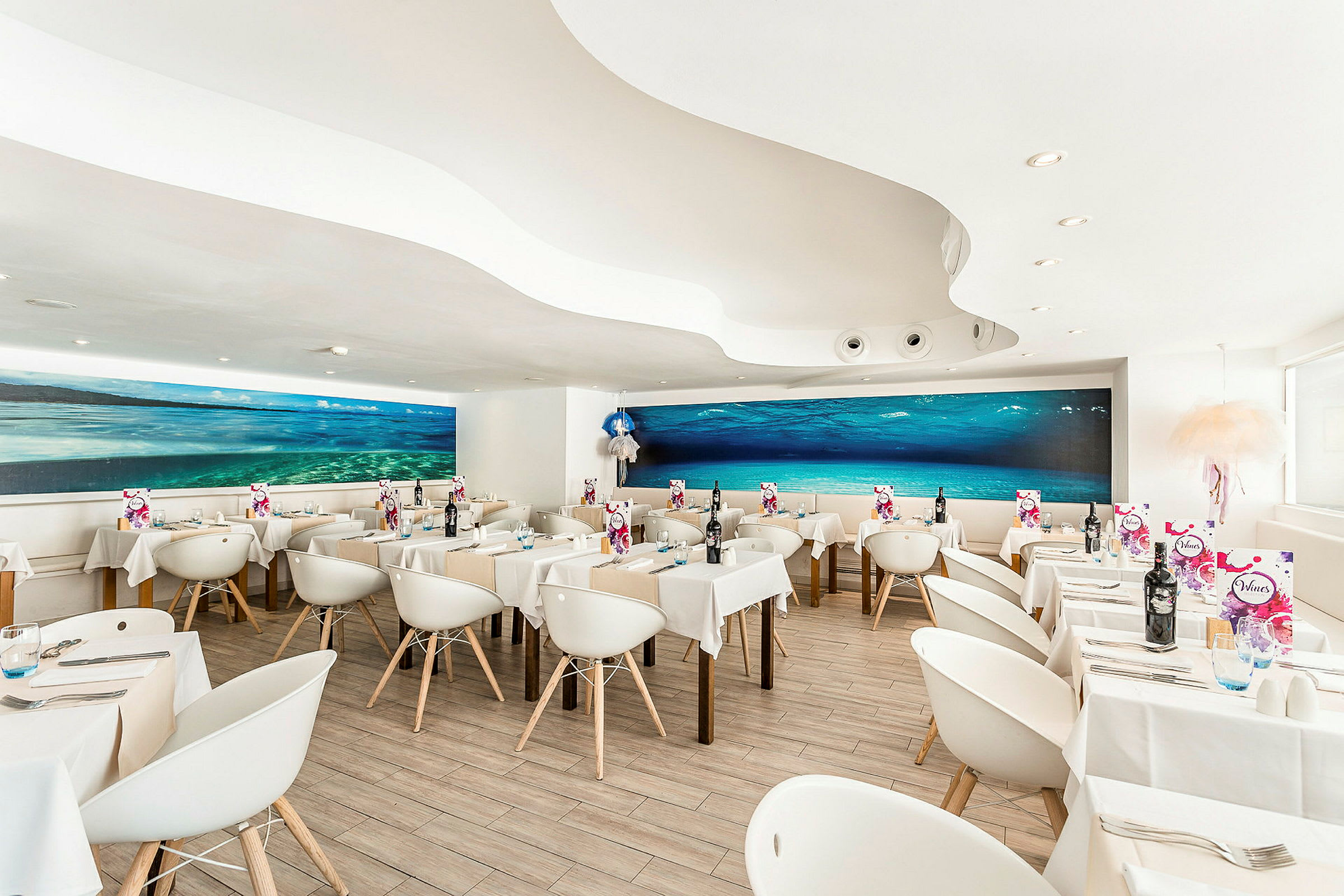 En moderne restaurant med hvite stoler og bord, samt et stort undervannsmaleri på veggen. (AI-generert tekst)