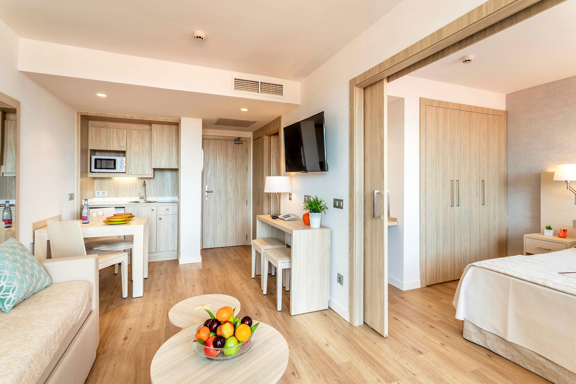 Moderne hotellrom med kjøkkenavdeling, bord, sofa og soverom. På sofabordet står en skål med fersk frukt. (AI-generert tekst)