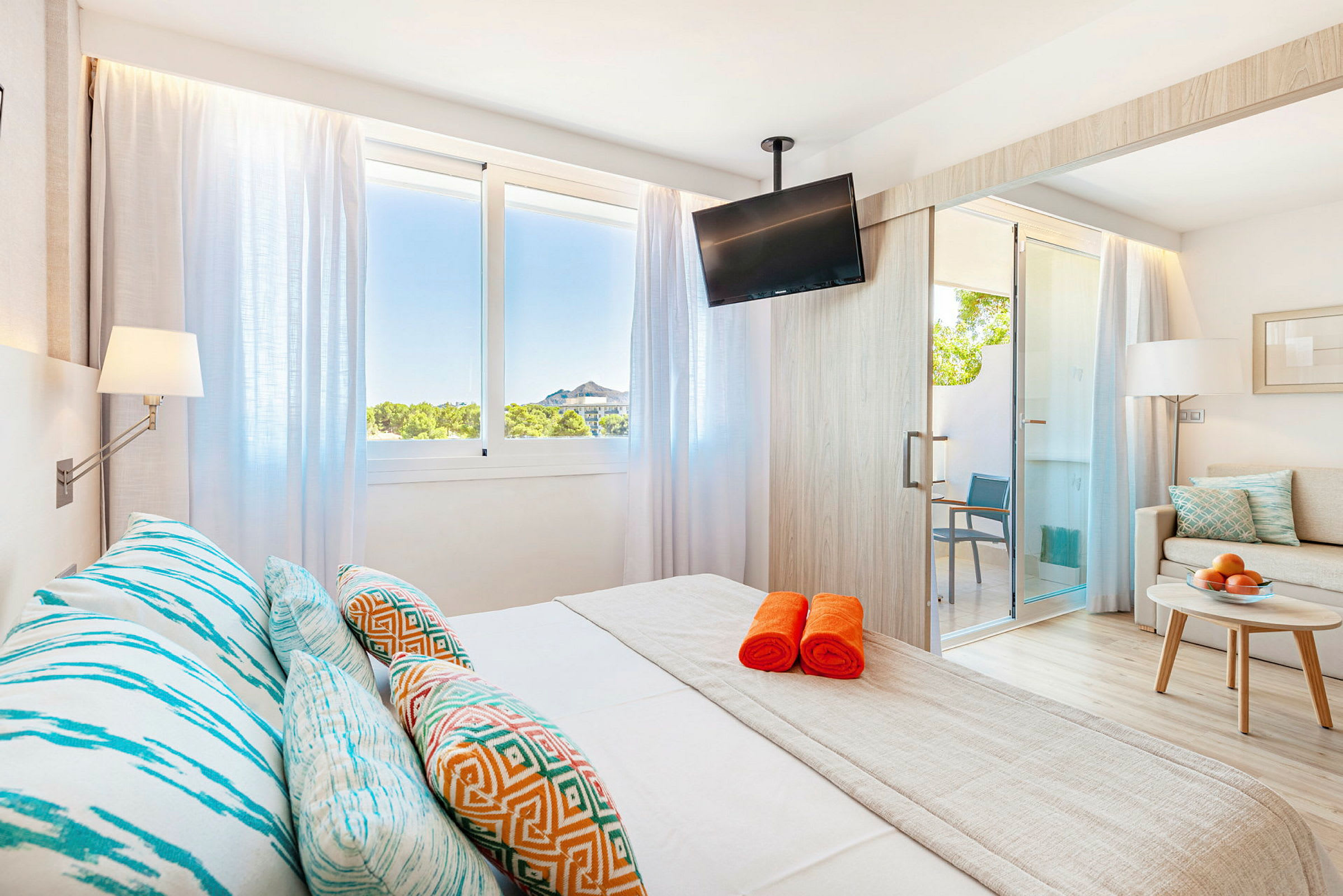 Lyst hotellrom med seng, fargerike puter og oransje håndklær. (AI-generert tekst)