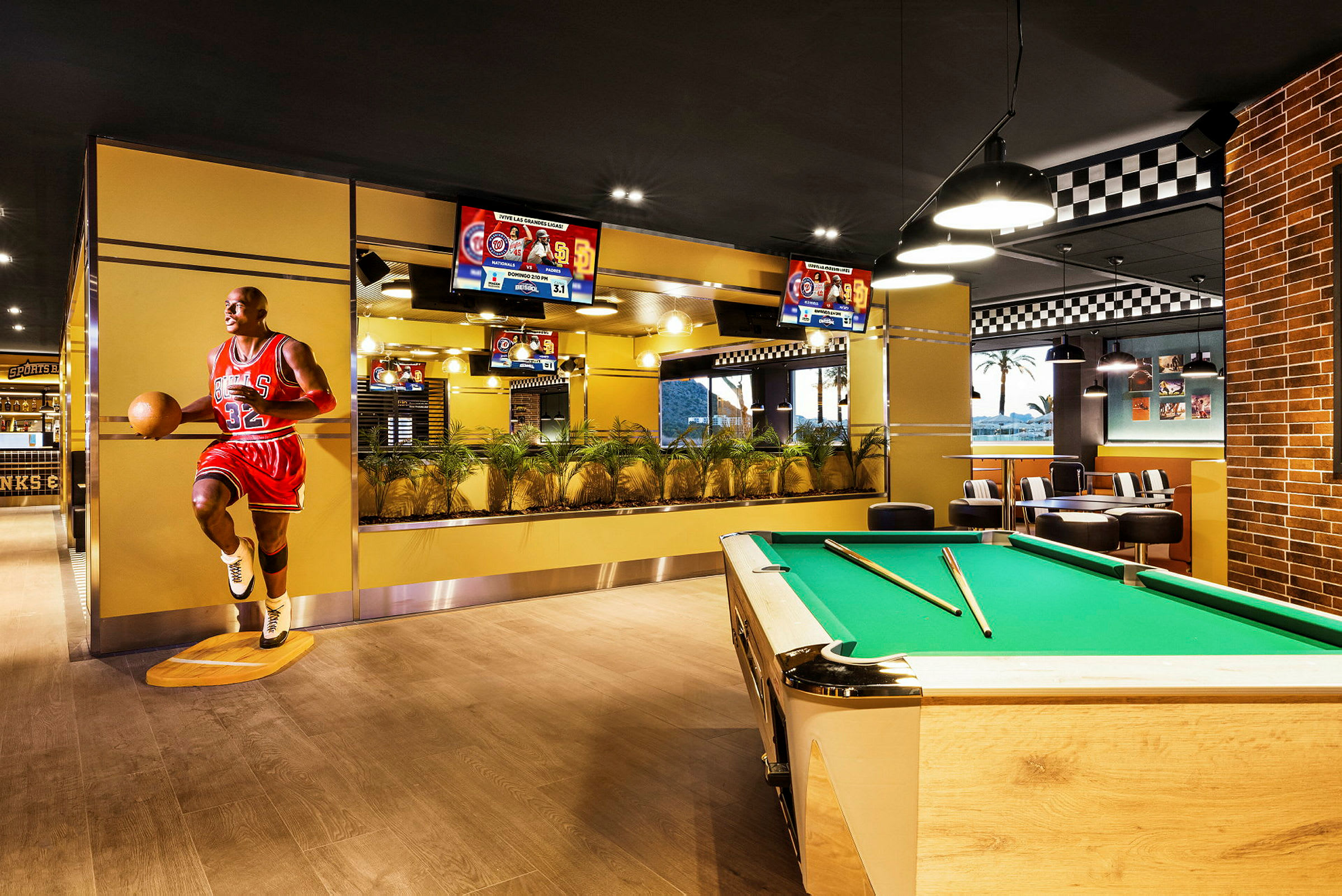 En innendørs scene med et biljardbord, en figur av en basketballspiller og flere skjermer som viser sportsbegivenheter. (AI-generert tekst)