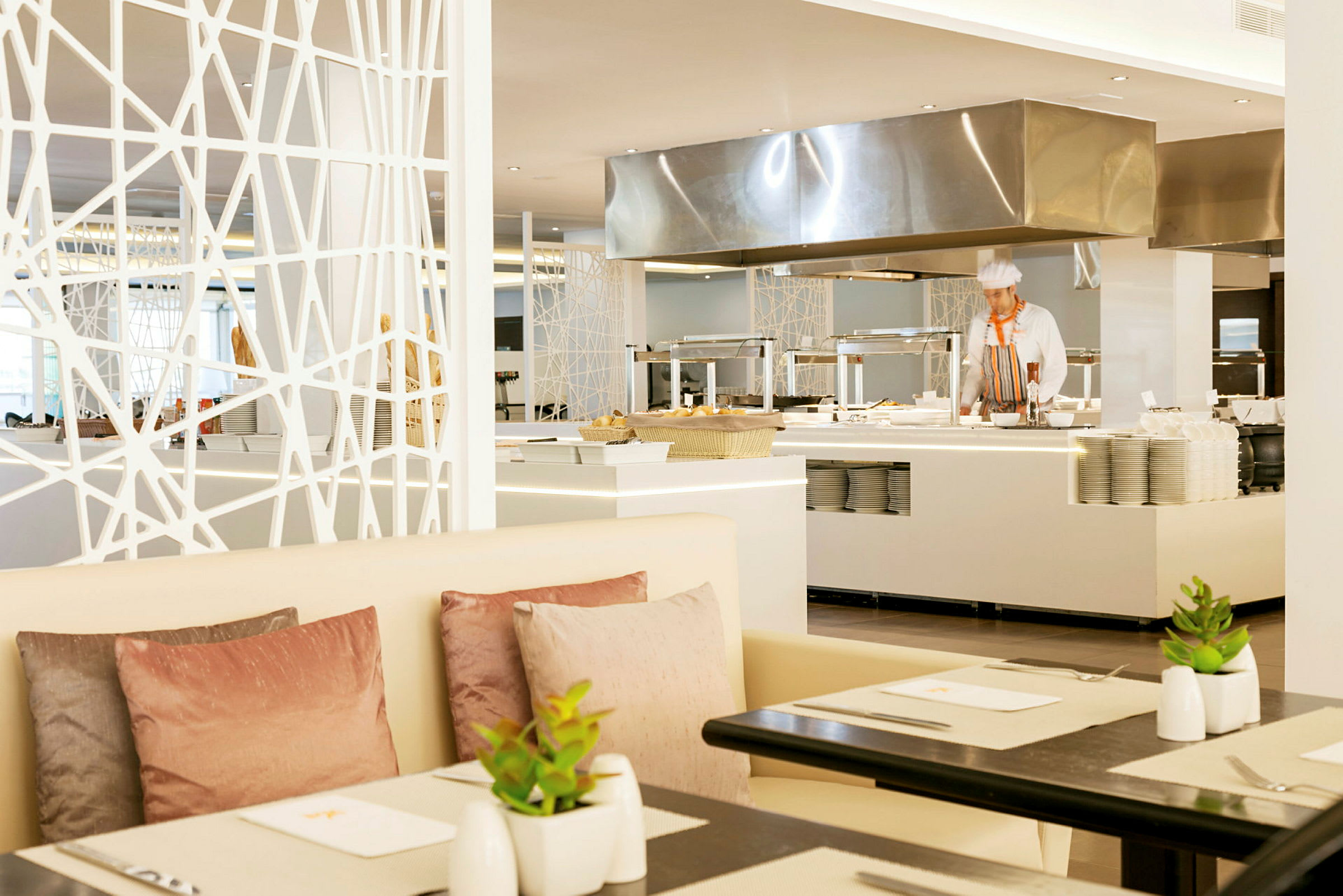 Et moderne restaurantområde med hvite møbler, pastellfargede puter og planter på bordene, i bakgrunnen er et åpent kjøkken synlig. (AI-generert tekst)