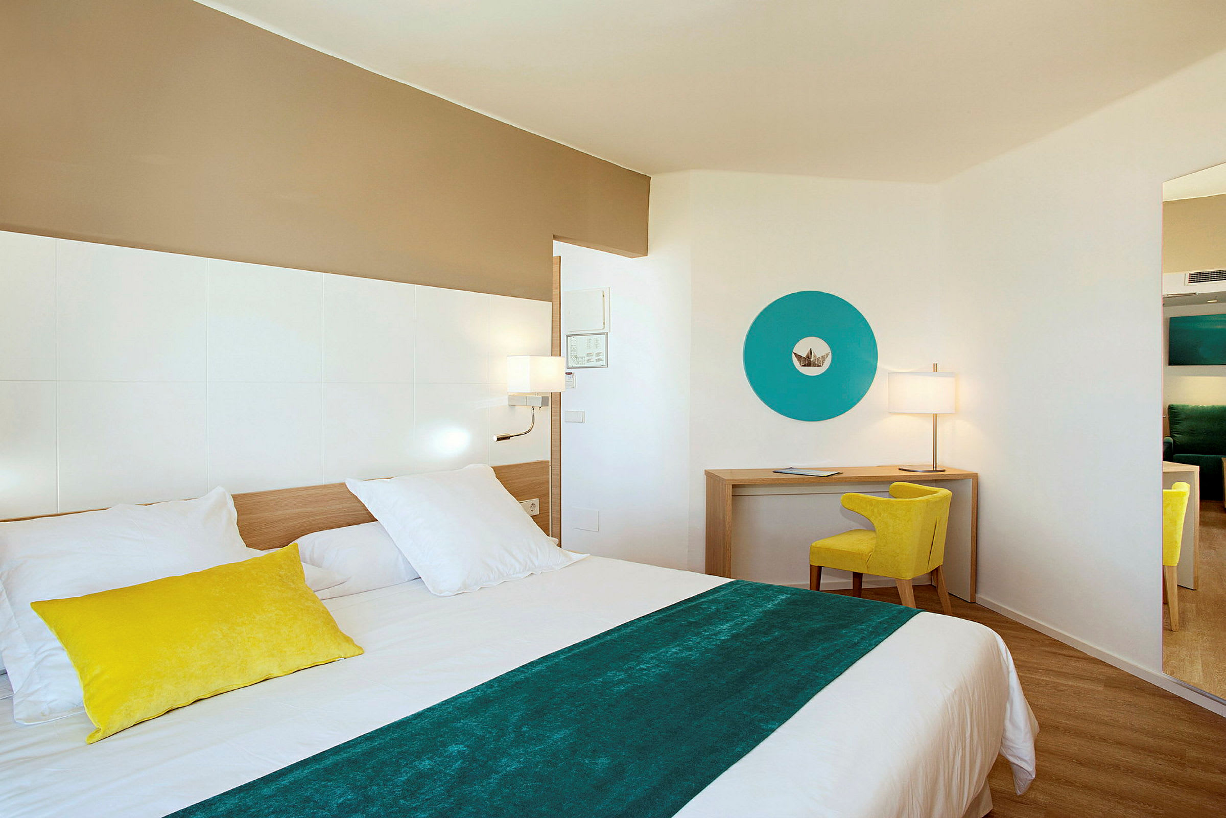Et moderne hotellrom med en seng i forgrunnen, et skrivebordområde med en gul stol og en blå dekorativ veggdetalj. (AI-generert tekst)