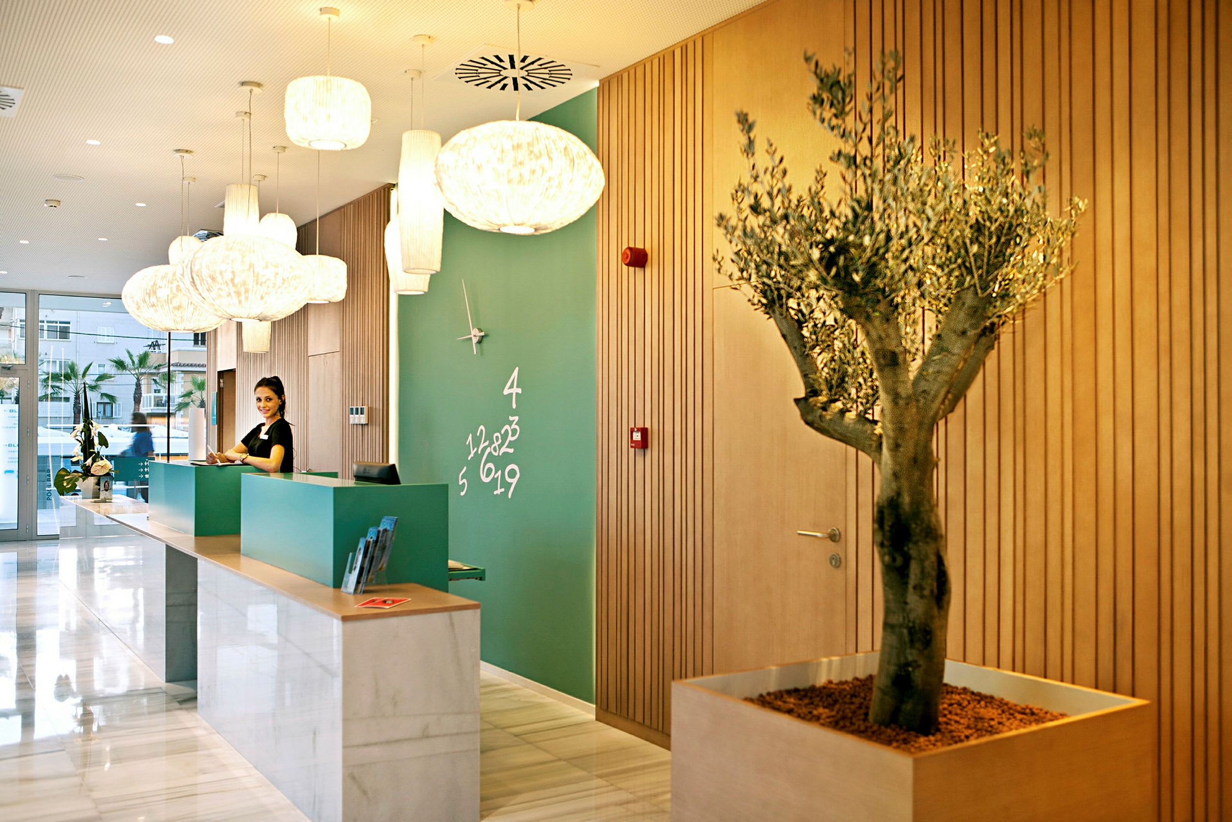 Resepsjonen på et hotell med moderne interiør, grønn vegg med en klokke og numerisk dekor, samt en trekrukke. (AI-generert tekst)