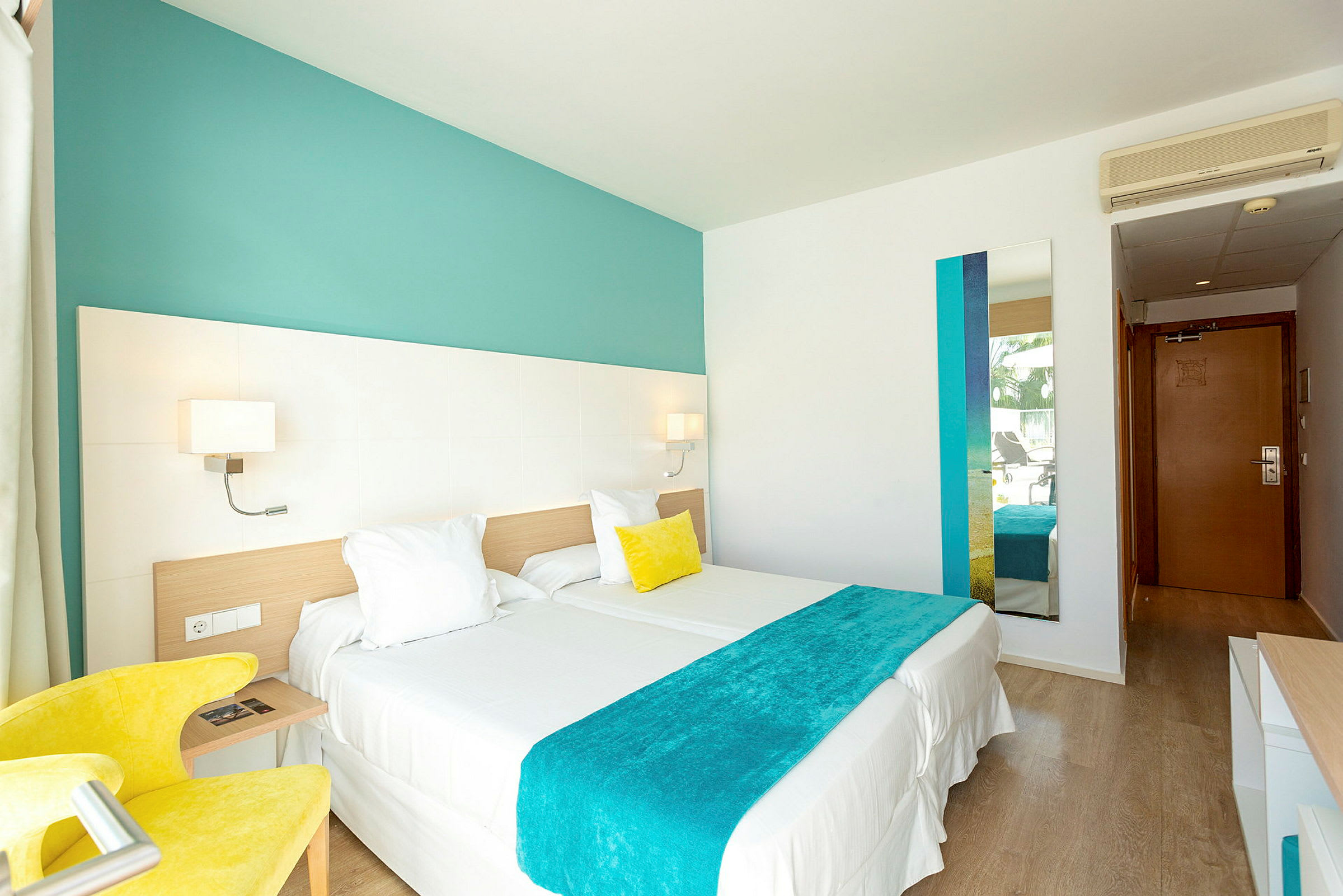 Et lyst hotellrom med to enkeltsenger som er satt sammen, en gul lenestol, veggdekor i turkise nyanser og et speil. En dør til inngangen er synlig i bakgrunnen. (AI-generert tekst)
