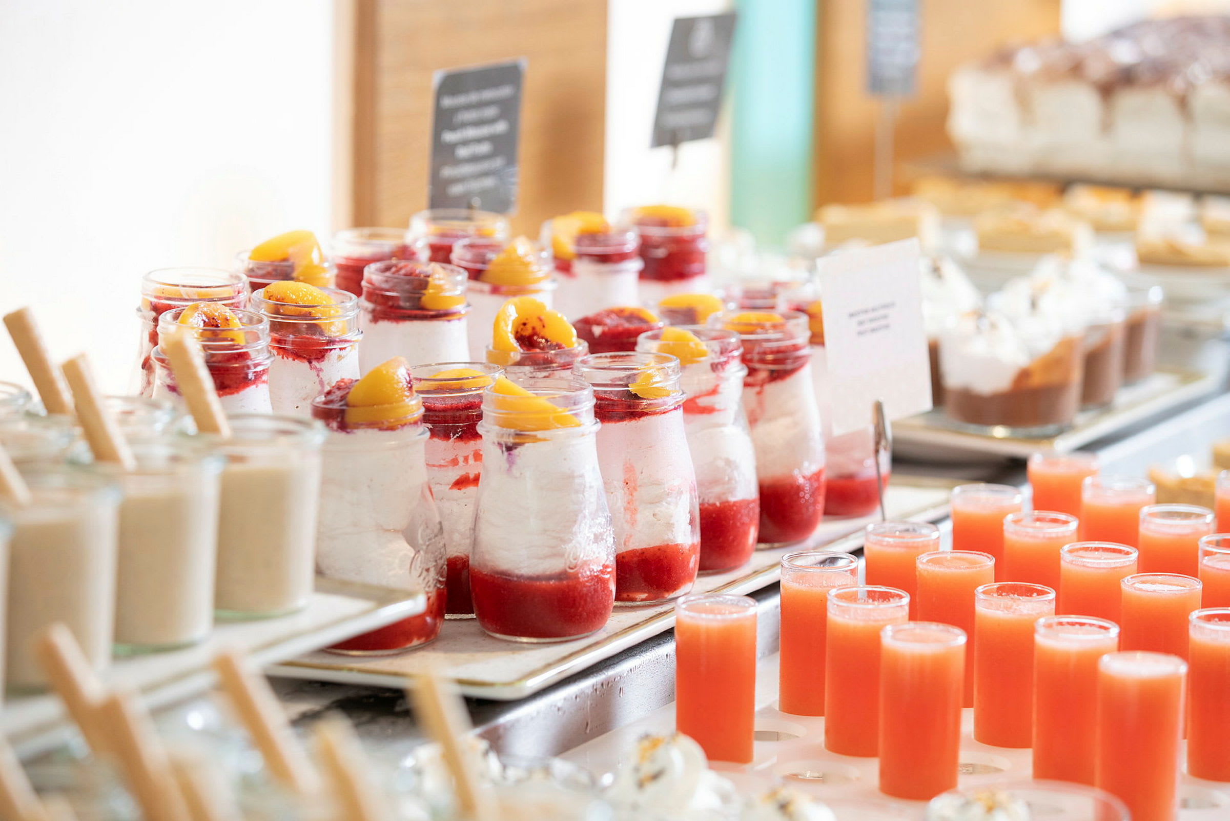 En rekke med desserter i glass med røde fruktdetaljer og små juiceglass på et bord. (AI-generert tekst)