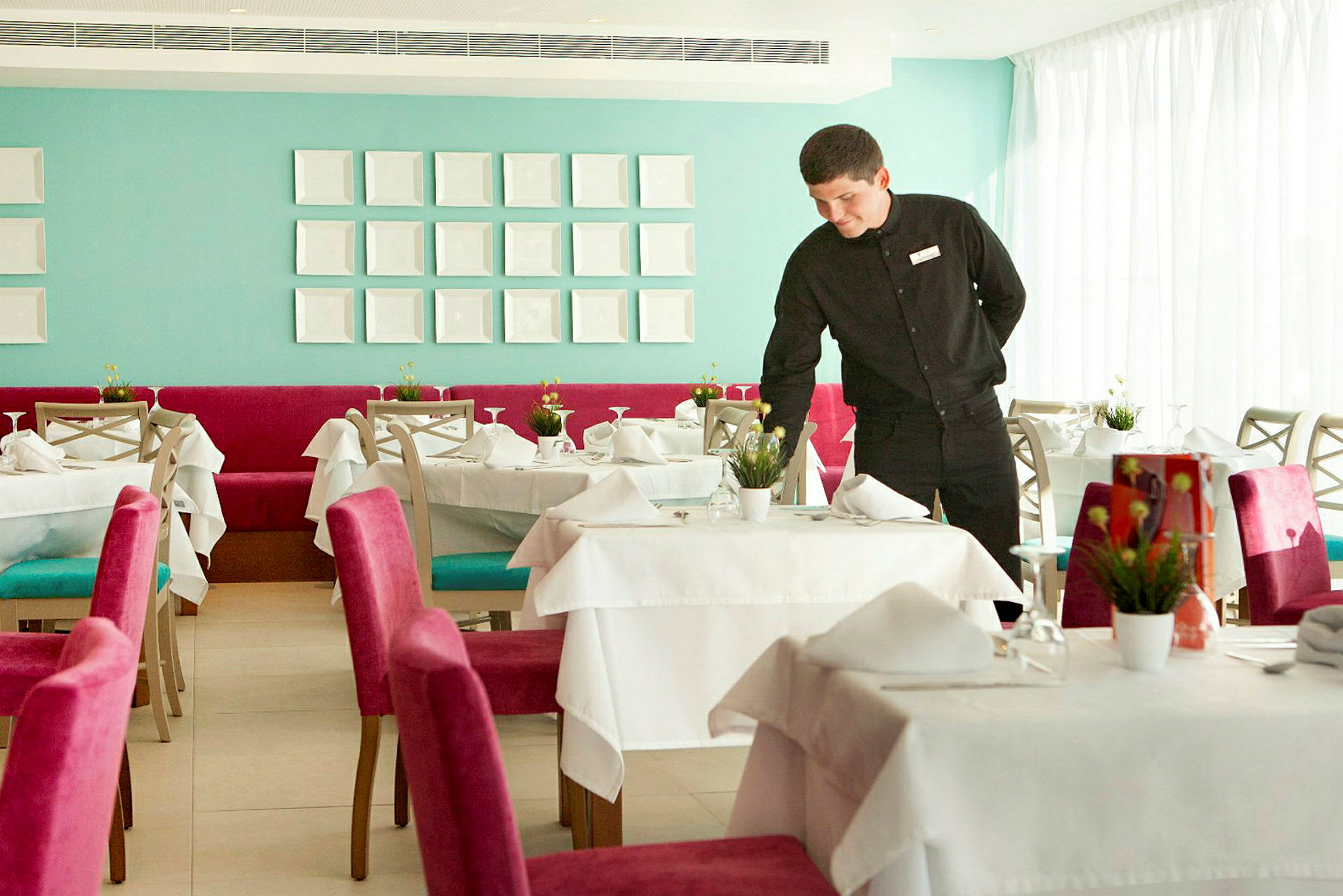En elegant restaurant med dekkede bord og fargerike stoler, en servitør forbereder et bord. (AI-generert tekst)