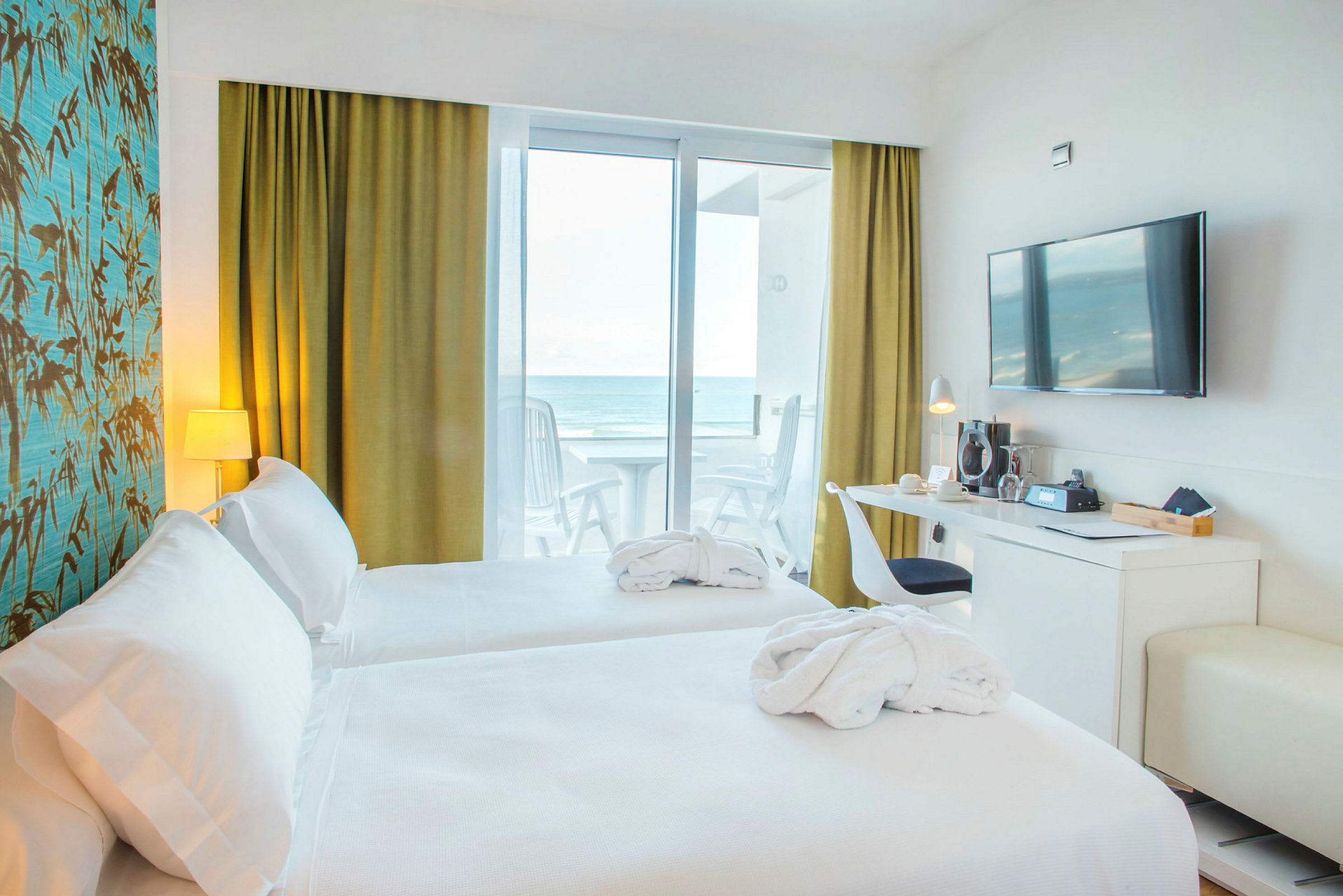 Hotellrom med to senger, skrivebord, vegg-TV og havutsikt fra balkongen. (AI-generert tekst)