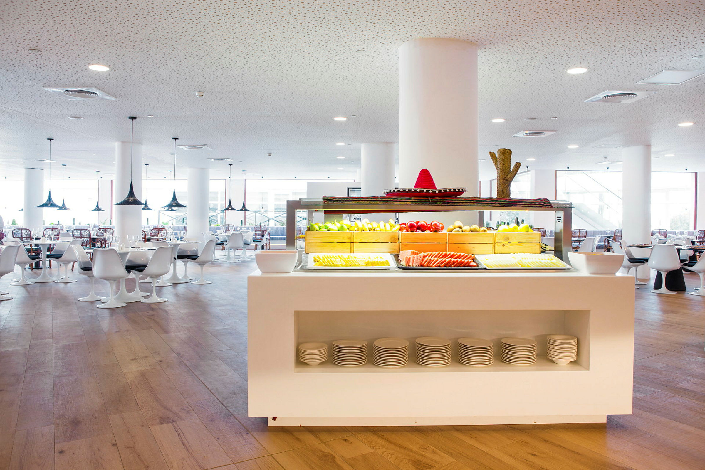 Et restaurantområde med en hvit bufféstasjon fylt med frisk frukt og grønnsaker, omgitt av moderne hvite bord og sittemøbler. (AI-generert tekst)