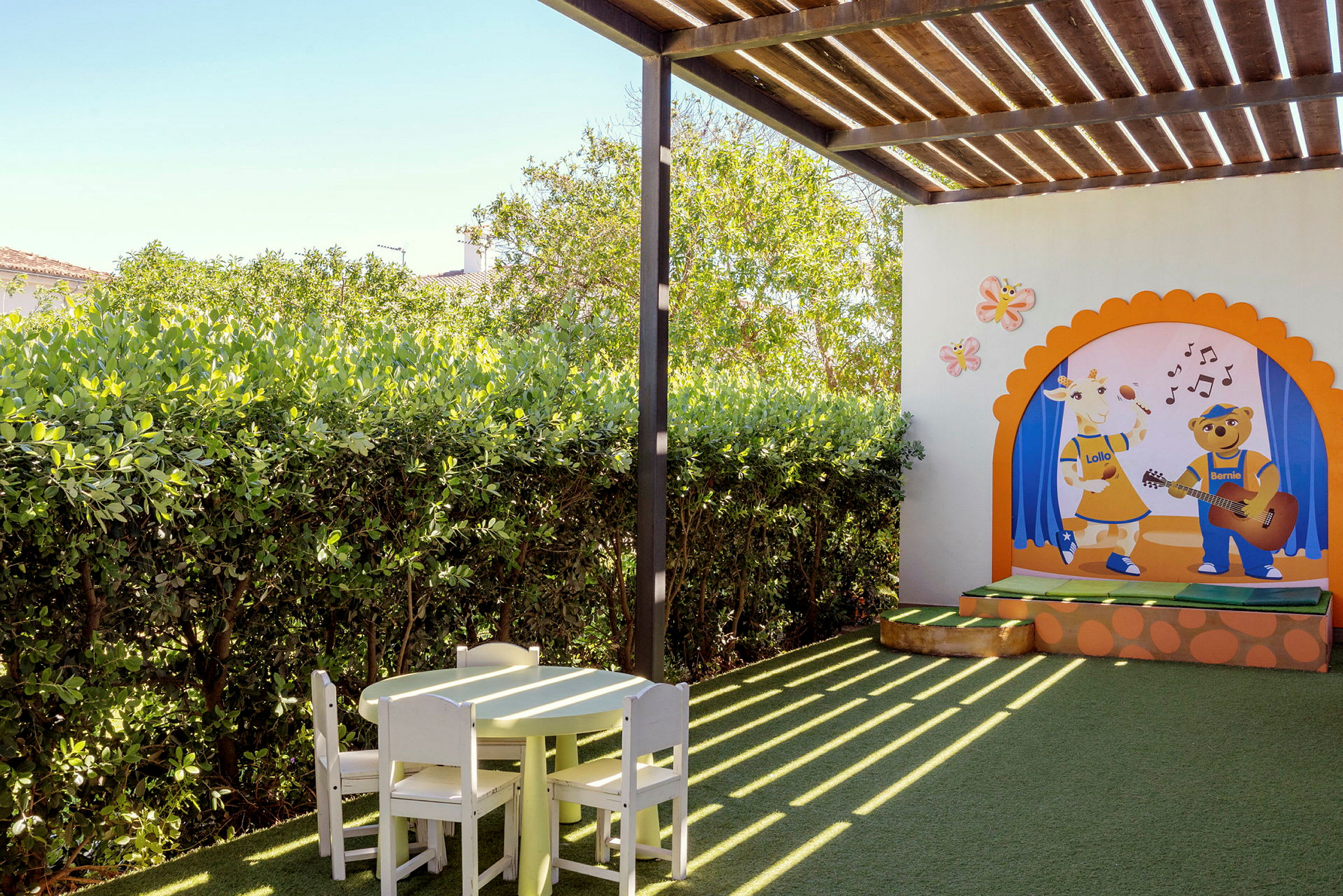 En utendørs lekeplass med et fargerikt veggmaleri som viser to figurer, samt et bord og stoler laget av tre under en trepergola. (AI-generert tekst)
