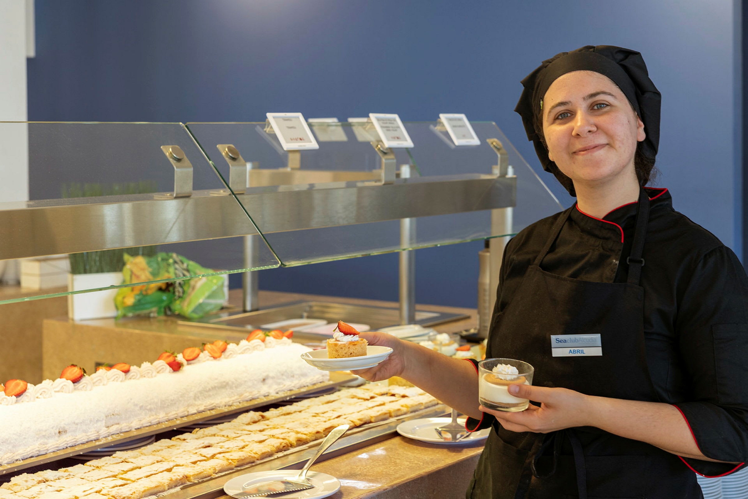 En kokk kledd i svart står ved en buffet med desserter og holder to porsjoner. (AI-generert tekst)
