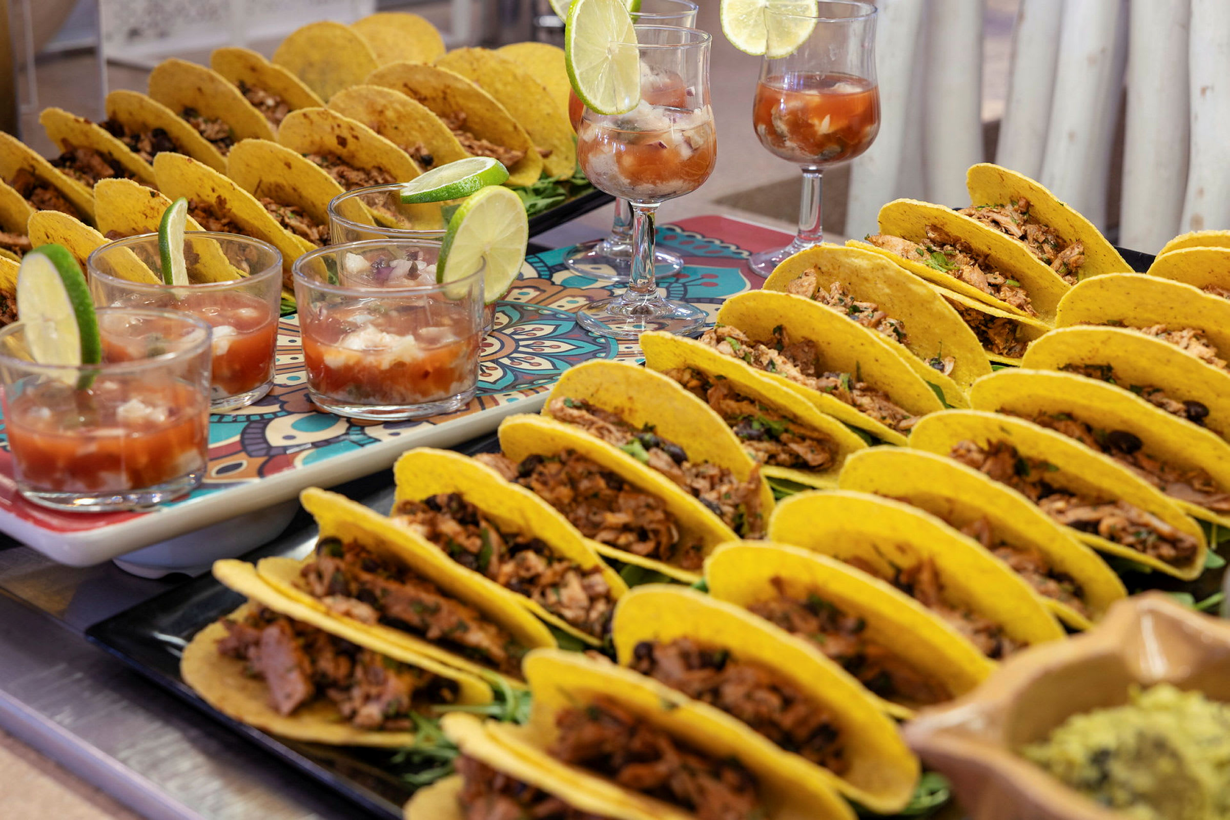 Bilde av flere tacos fylt med kjøtt og grønnsaker, servert på fat sammen med glass med drikke og limebåter. (AI-generert tekst)
