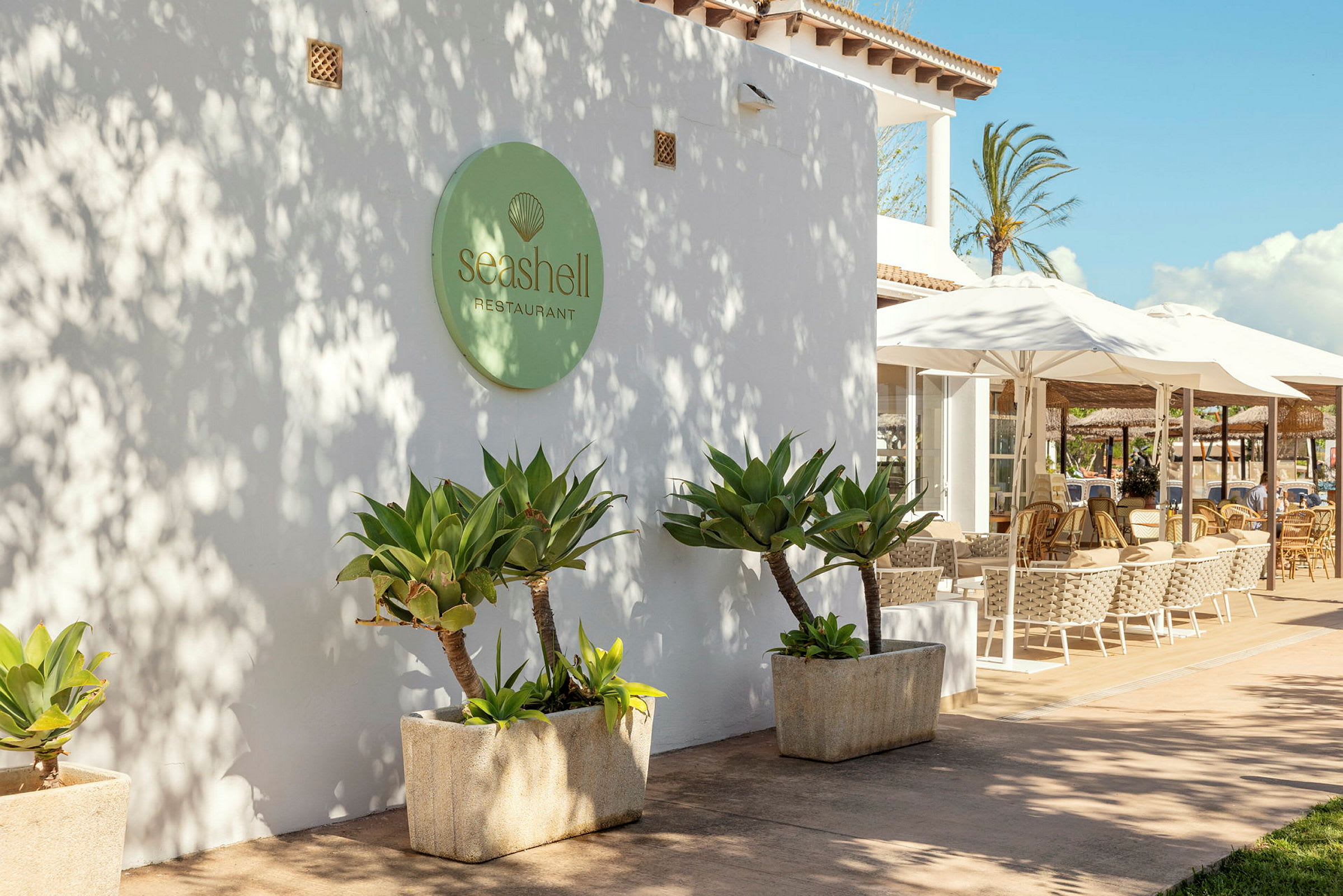 Utvendig av restauranten Seashell med hvite vegger, grønne planter og uteservering med paraplyer. (AI-generert tekst)