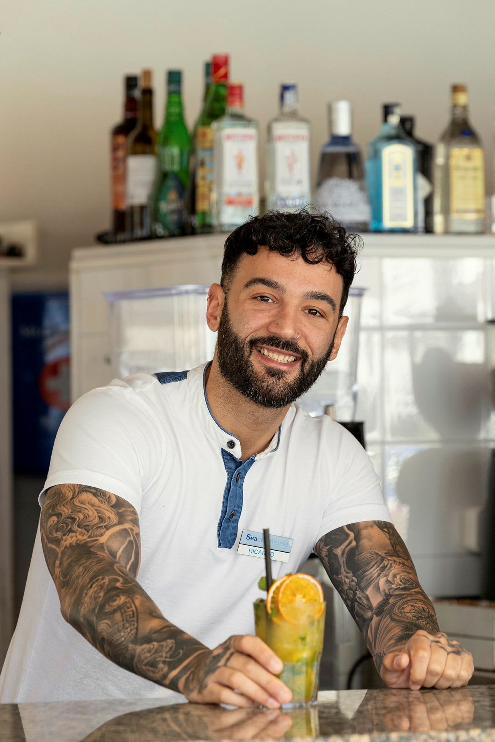 En smilende mann står ved en bardisk med forskjellige spritflasker i bakgrunnen og holder en cocktail med appelsinskive. (AI-generert tekst)