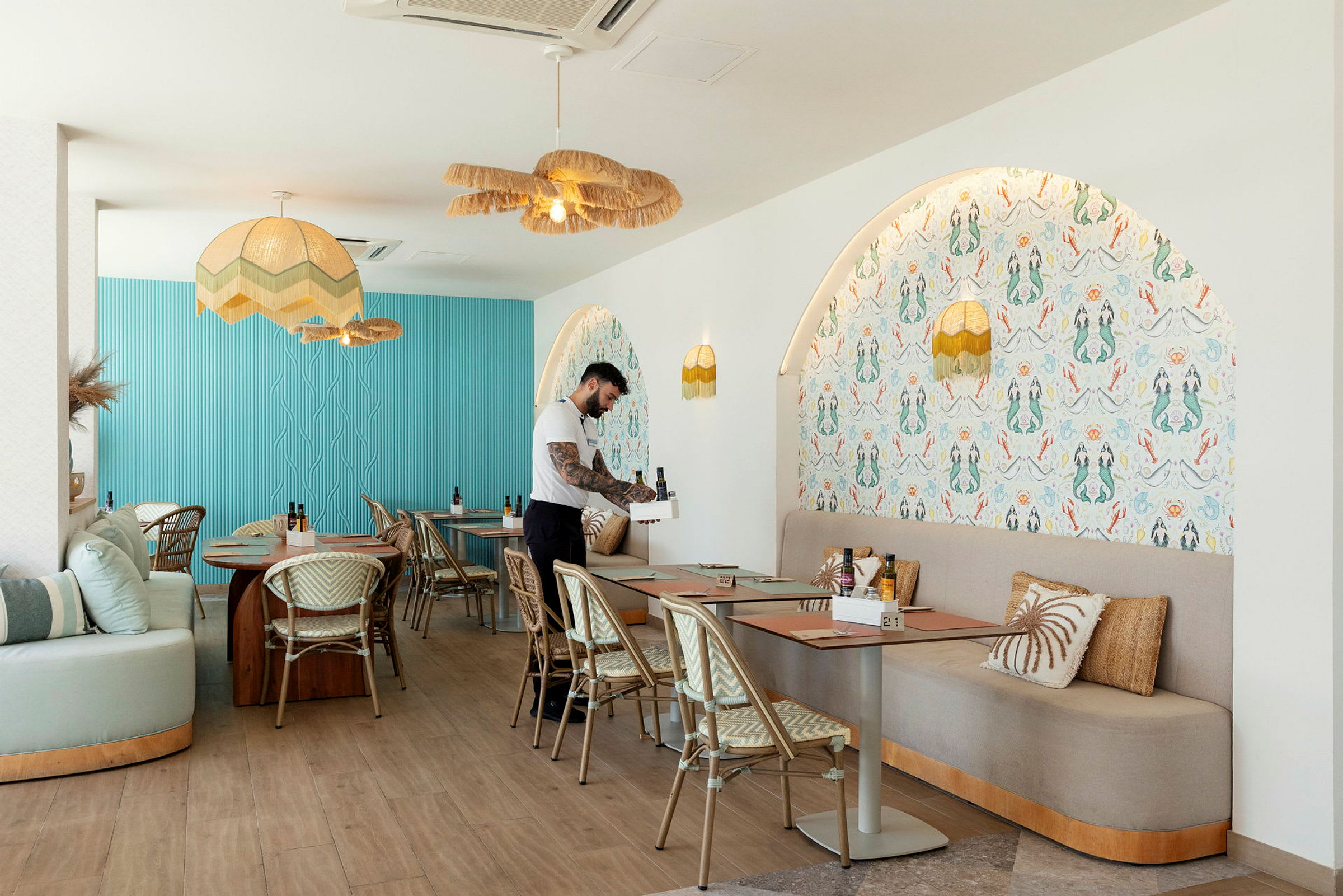 En interiør av en moderne restaurant med blå og mønstrede vegger, bord med stoler og en servitør som forbereder en plass. (AI-generert tekst)