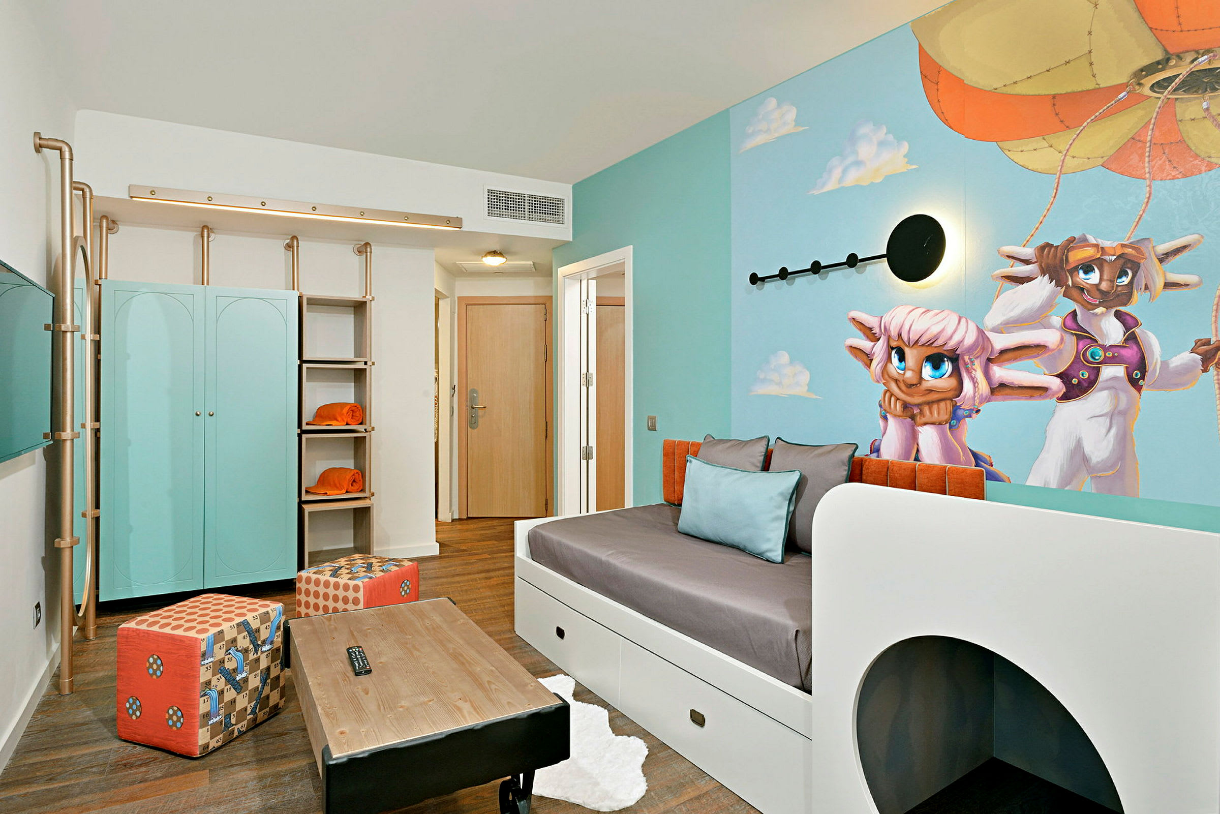 Et hotellrom med en lys og fargerik innredning, inkludert et blått skap, oransje detaljer og en veggmaleri av to figurer foran en luftballong. (AI-generert tekst)