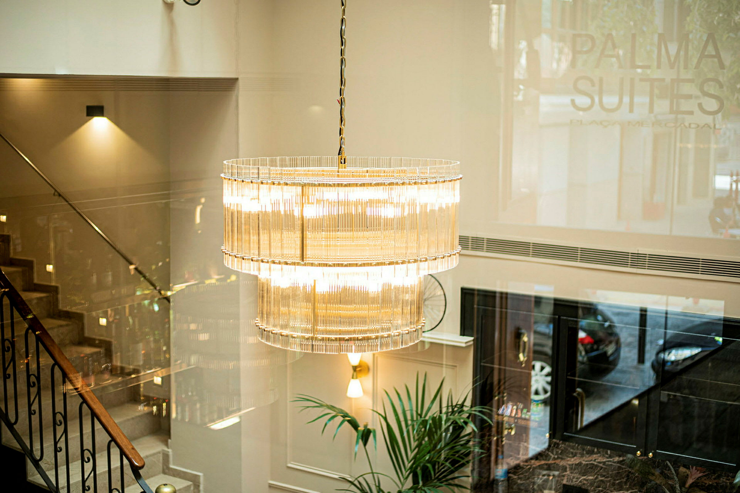 En elegant lysekrone henger i en lobby med trapp og plante synlig. (AI-generert tekst)